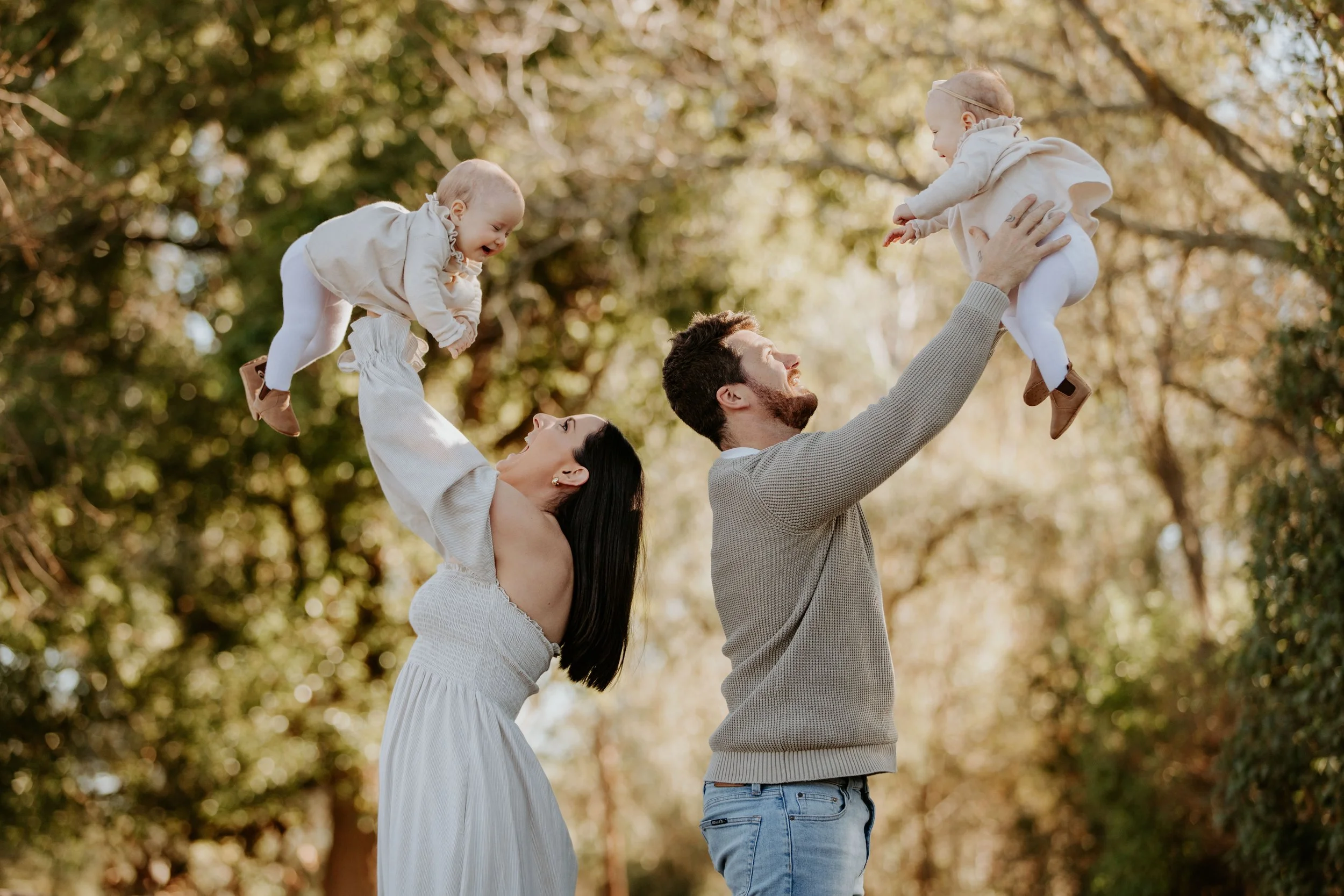 A family of four outdoors surrounded by trees, with two parents lifting their young children in the air, both children smiling and reaching towards each other.