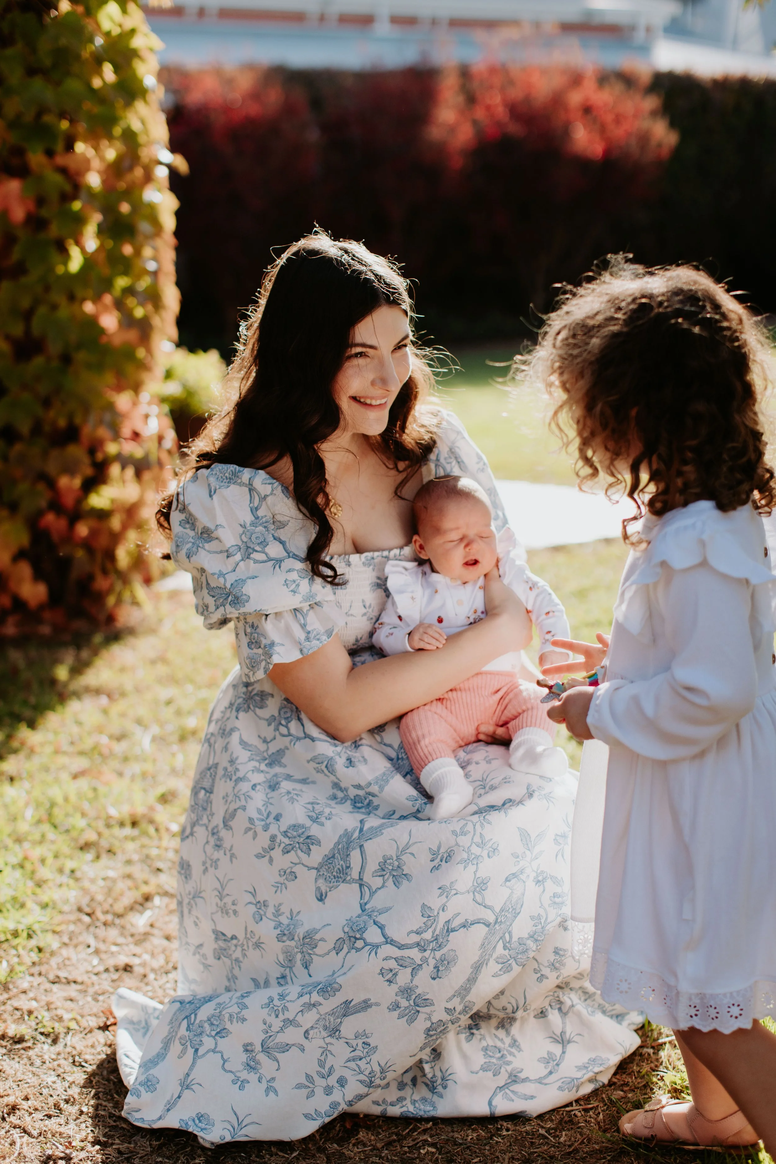 A woman with long dark hair in a floral dress kneels outdoors, holding a crying baby girl dressed in a white top and pink shorts. A young girl with curly hair in a white dress stands nearby, interacting with the woman.