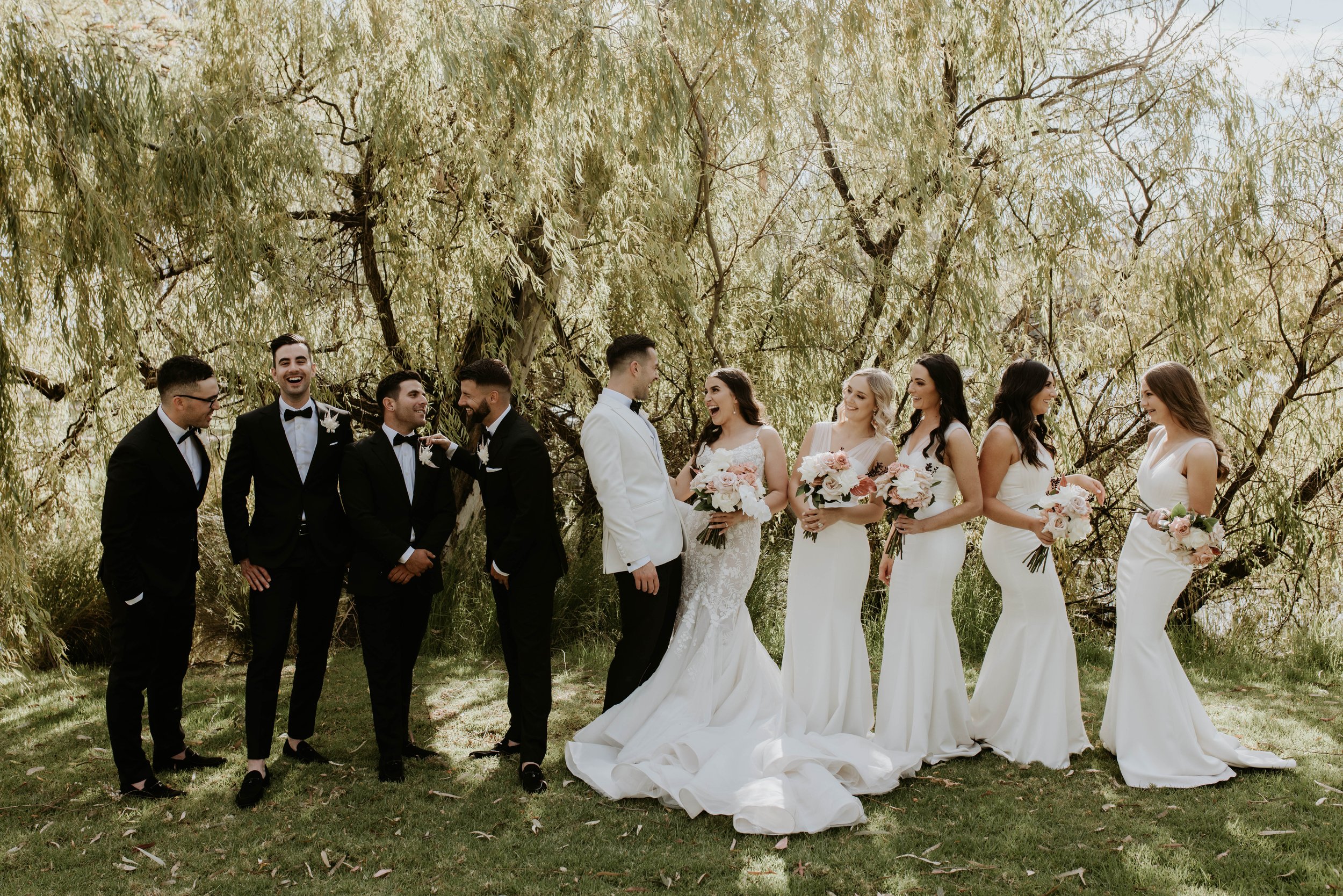 Bride and groom with their wedding party outdoors under trees, with the bridesmaids in white dresses holding bouquets and the groomsmen in black suits with bow ties.