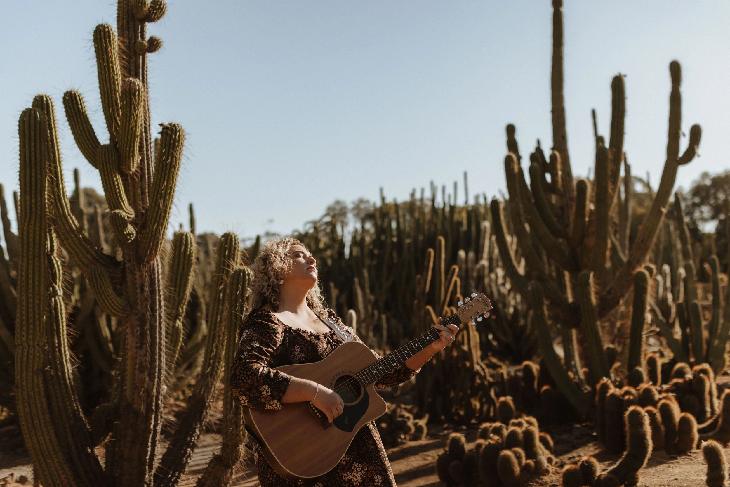 A woman with curly blonde hair standing among tall cacti, playing an acoustic guitar during daytime. Cactus Country, Strathmerton