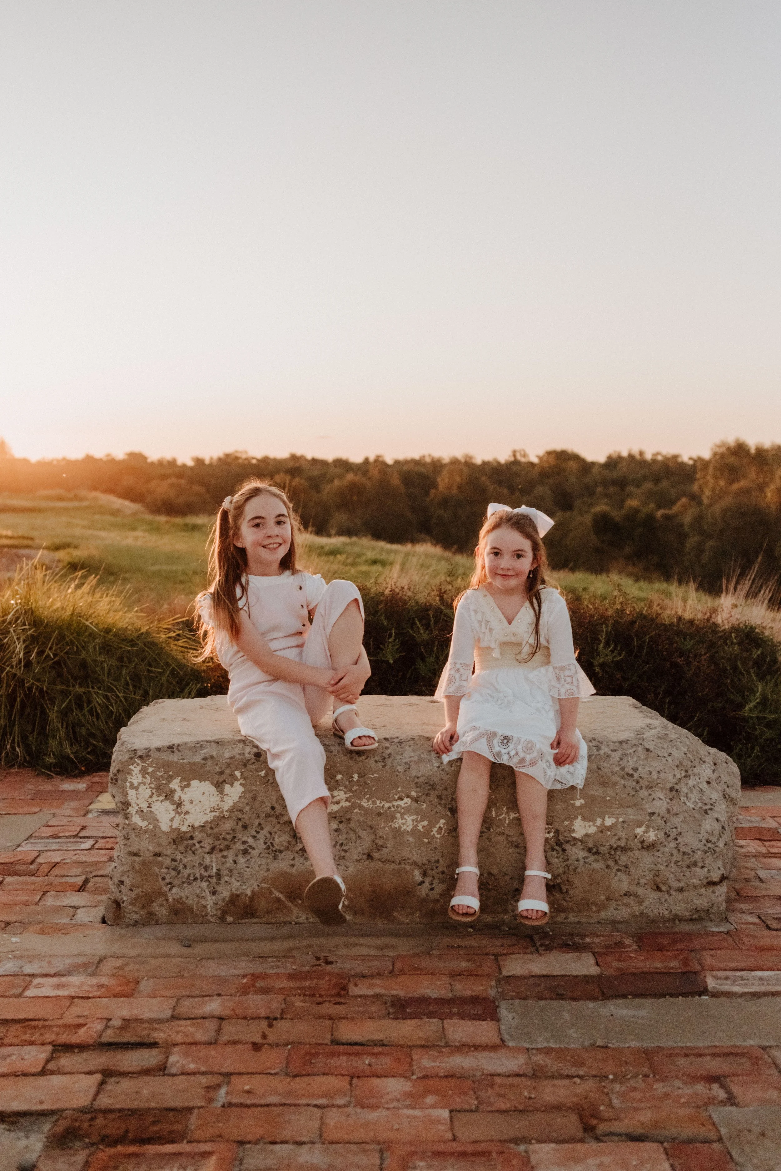 Two young girls sitting on a large rock outdoors during sunset, wearing white dresses and sandals, with a natural landscape and trees in the background.