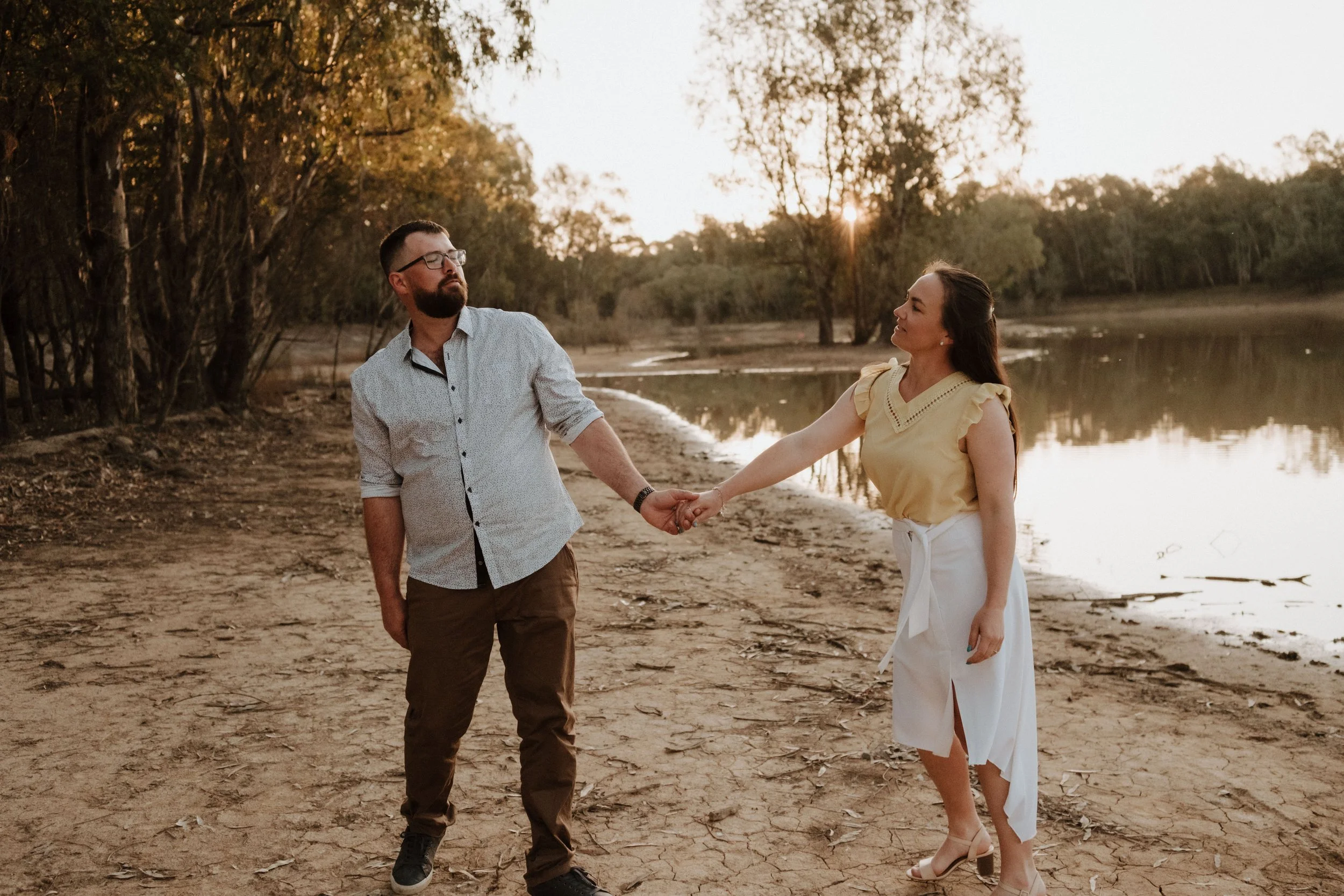 A man and woman holding hands on a sandy lakeshore during sunset, surrounded by trees and calm water.