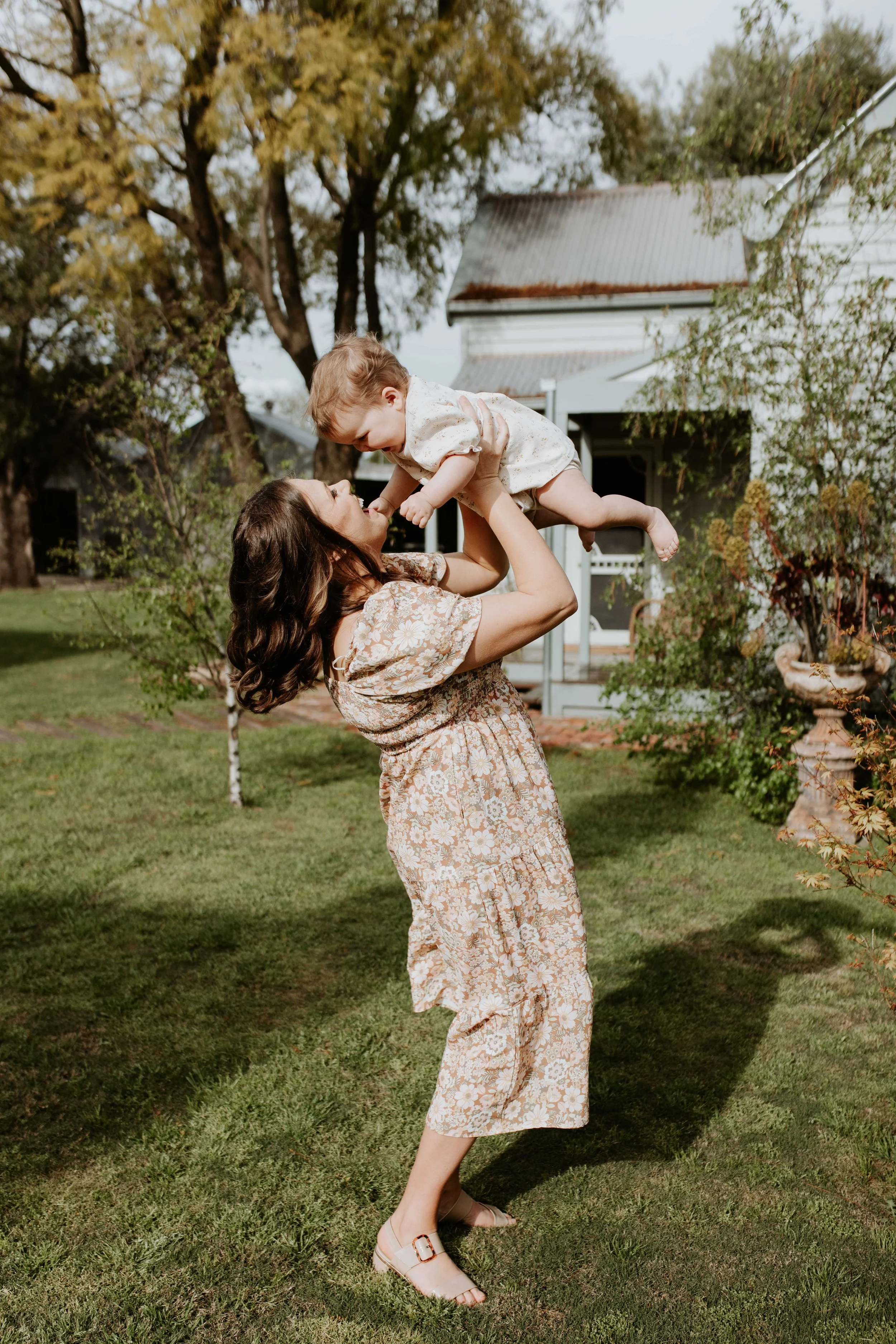 A woman lifting a young child in the air outdoors on a grassy yard with trees and a white house in the background.