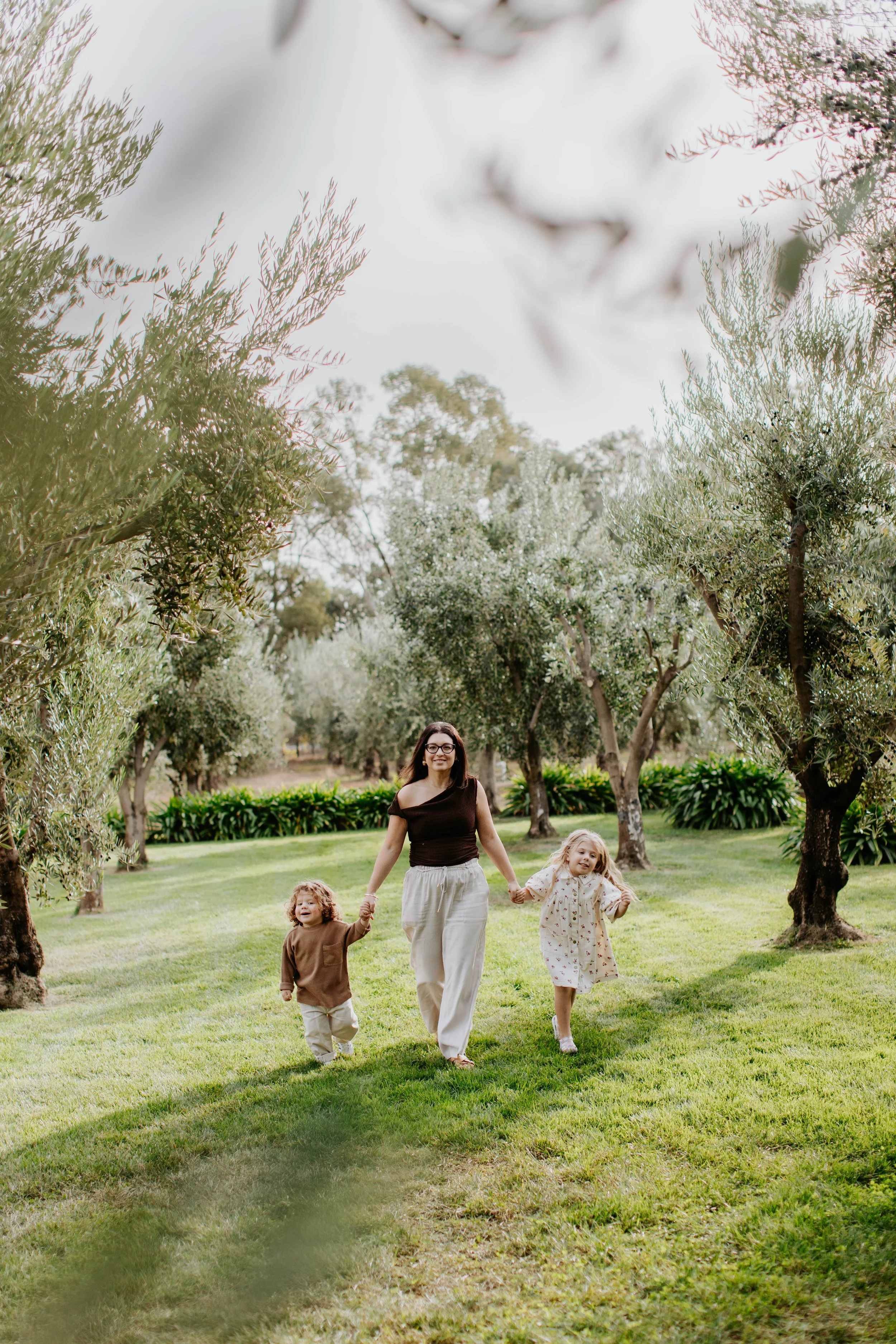 A woman and two children holding hands and walking through a green park with trees and grass - Olivehouse, Kialla.
