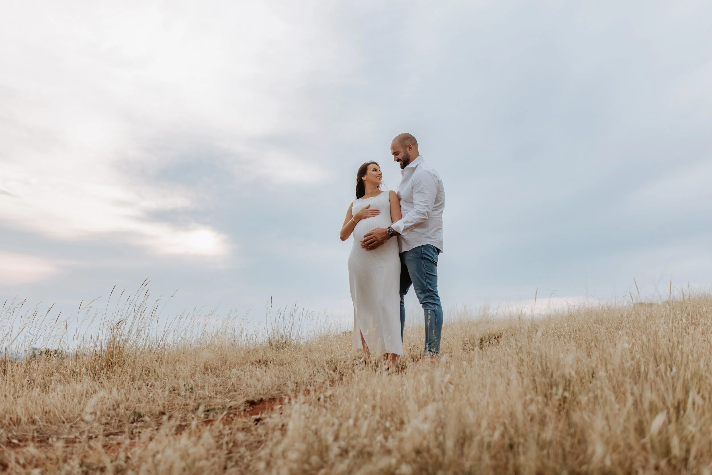 A pregnant woman and a man standing in a grassy field, smiling and touching her belly, under a cloudy sky.