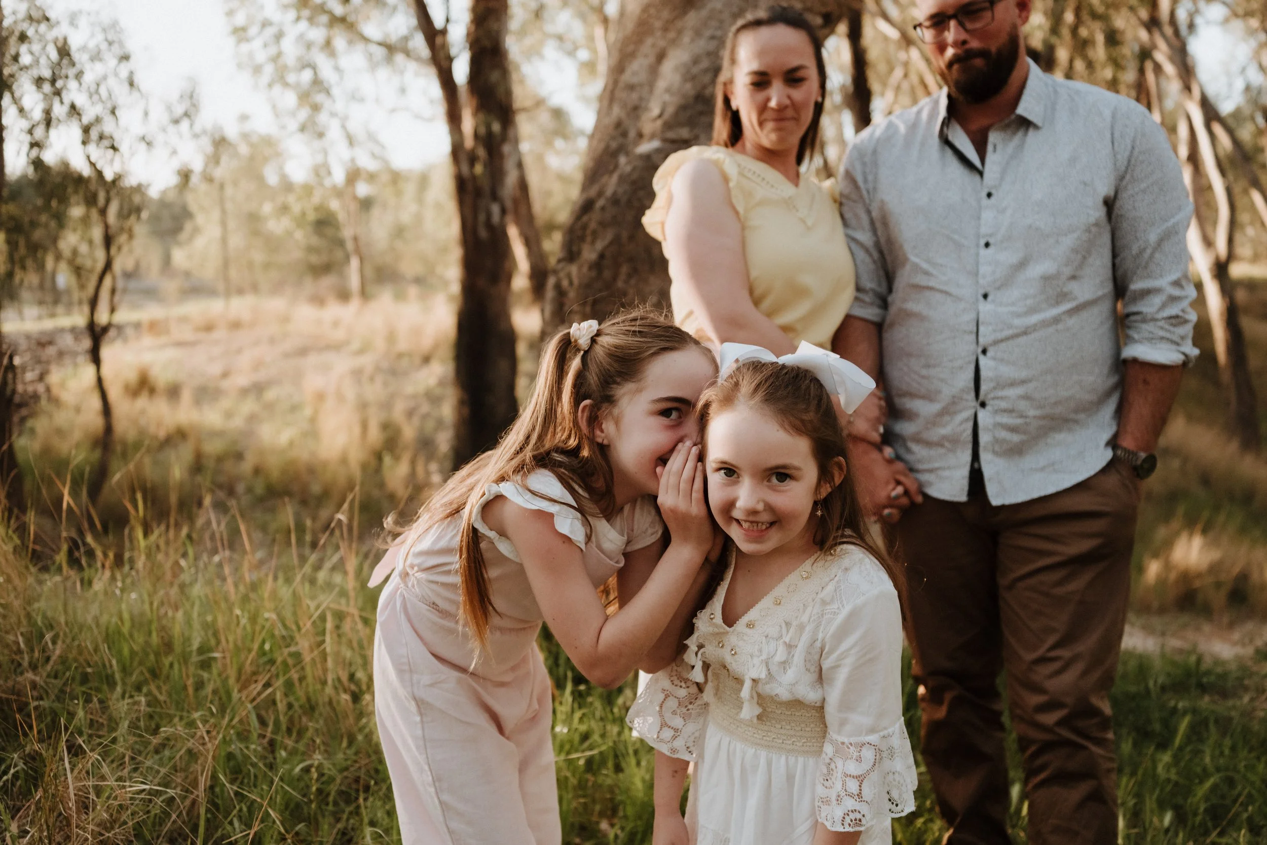 A family of four, two adults and two young girls, standing outdoors in a grassy area with trees. The girls are smiling and whispering to each other, wearing light-colored dresses. The adults are behind them, holding hands, and smiling.