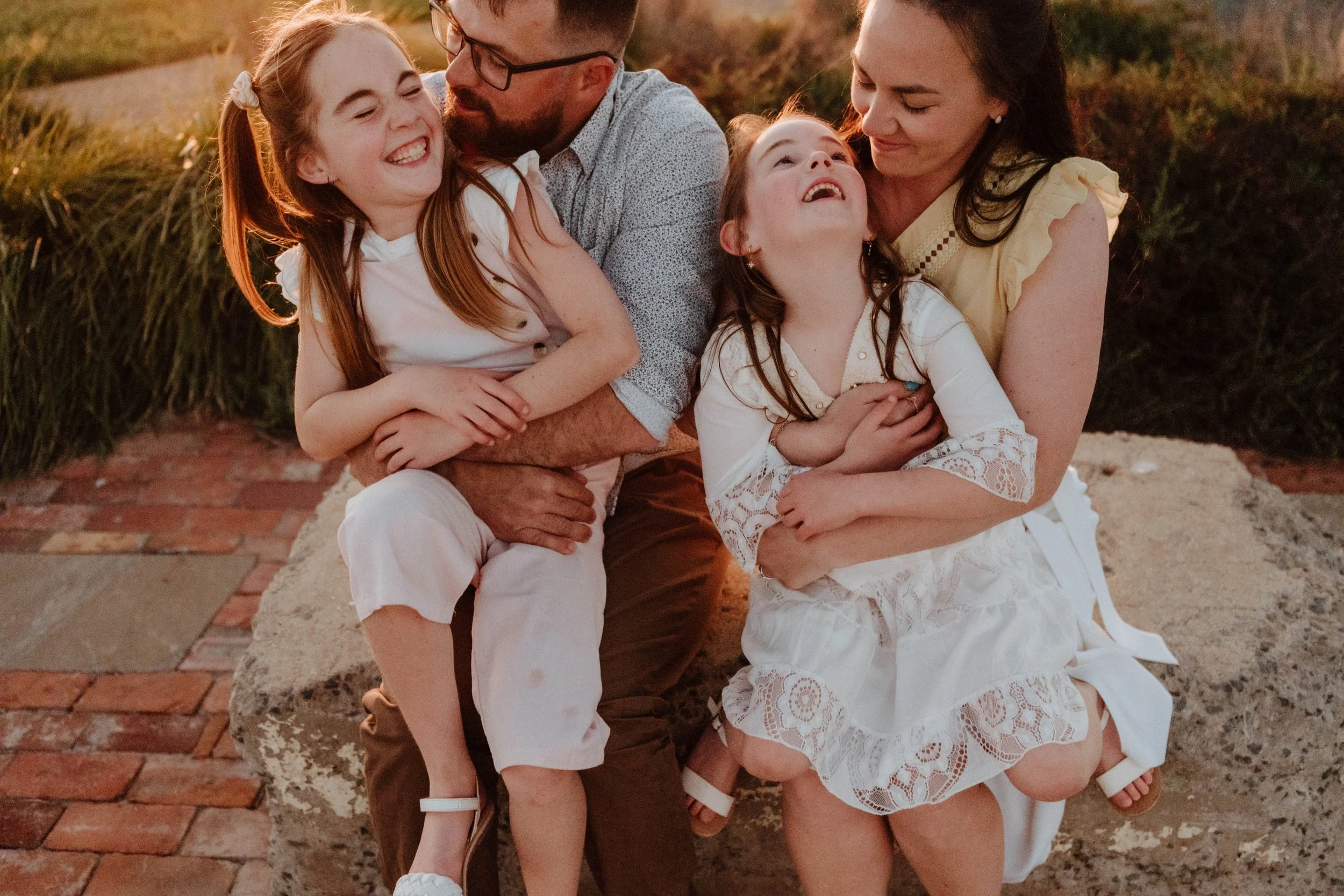 A family of four sitting on a stone bench outdoors, smiling and laughing together during a sunny day.