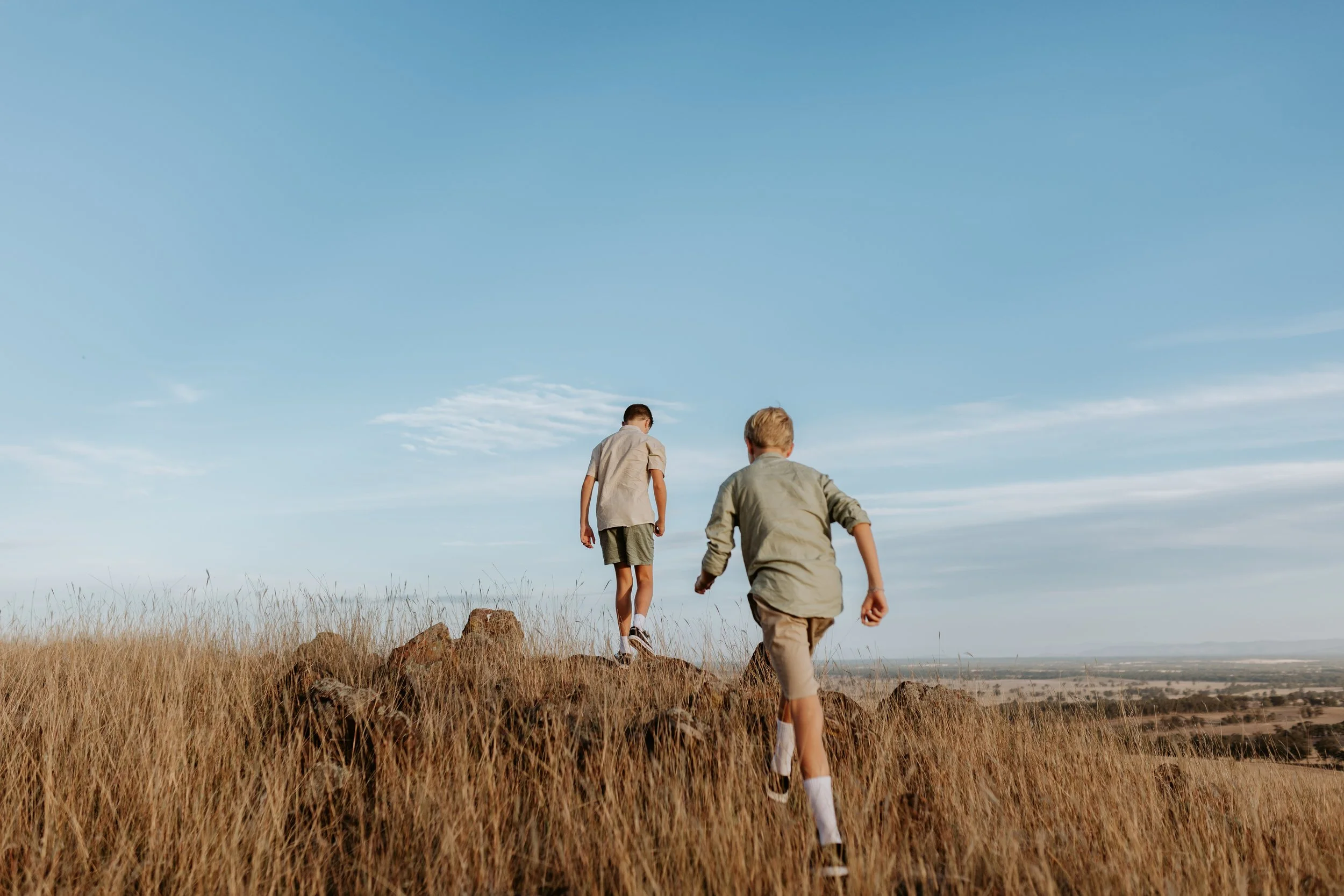 Two boys hiking on a dry, grassy hill under a blue sky, with a distant landscape visible in the background.