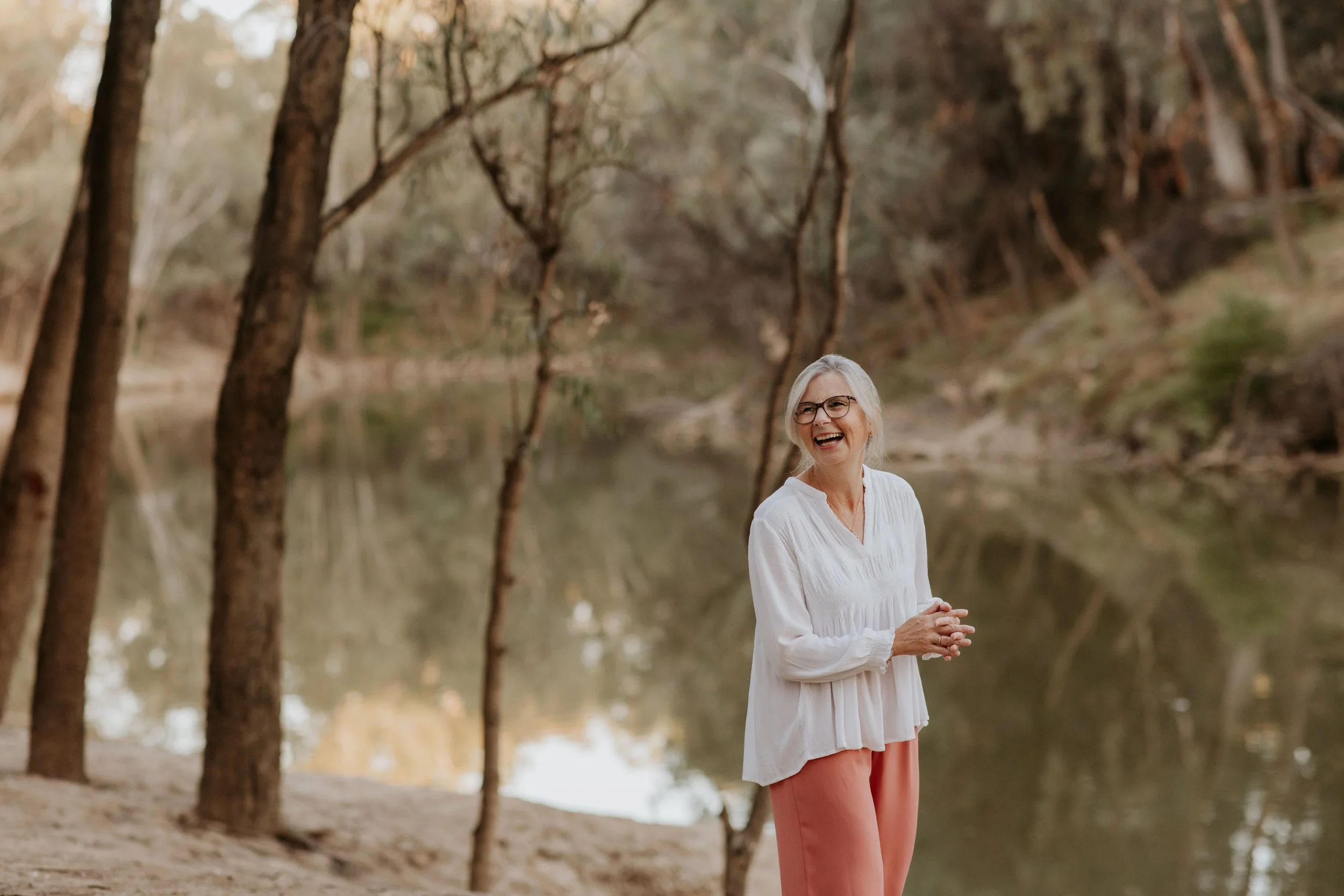 An elderly woman with gray hair, glasses, wearing a white blouse and peach pants, standing by a river with trees in the background, smiling and laughing.