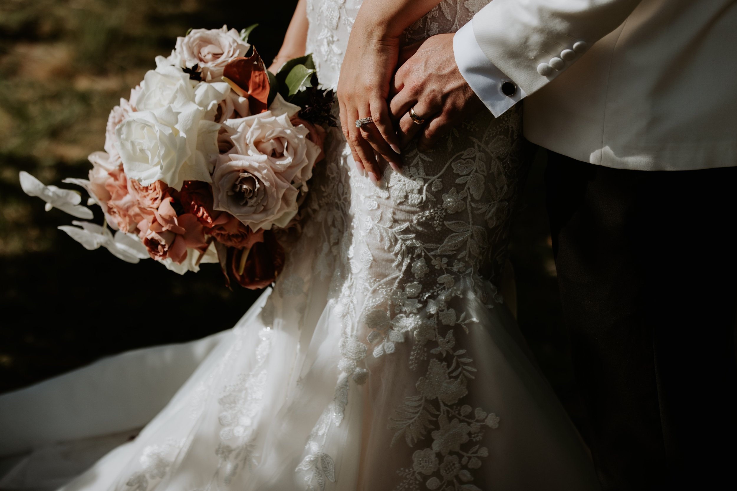 Close-up of a bride and groom holding hands, bride wearing a lace wedding dress, groom in a white suit jacket, holding a bouquet of pastel-colored roses and flowers.