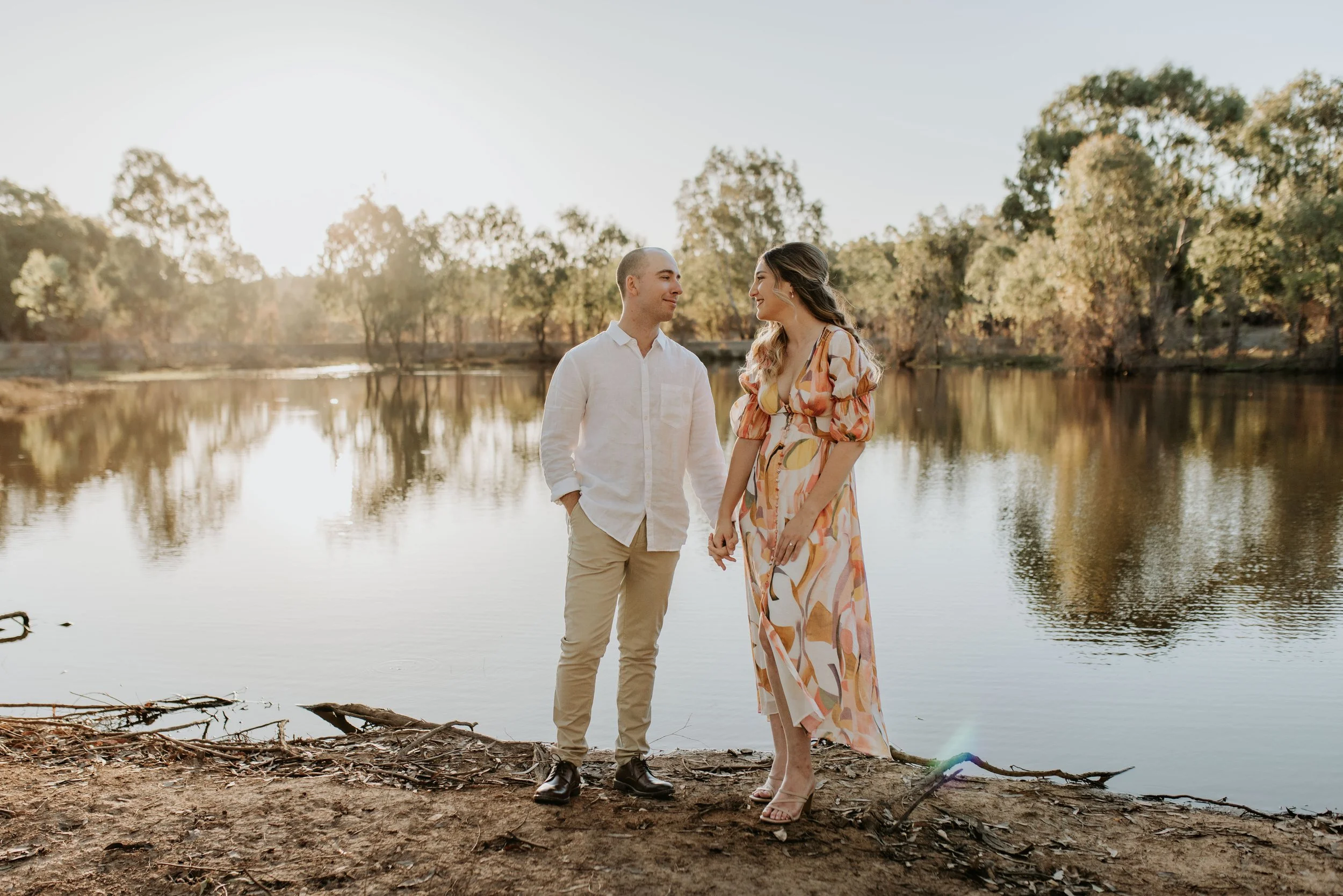 A man and woman holding hands by a lake during sunset, smiling and looking at each other, with trees in the background.