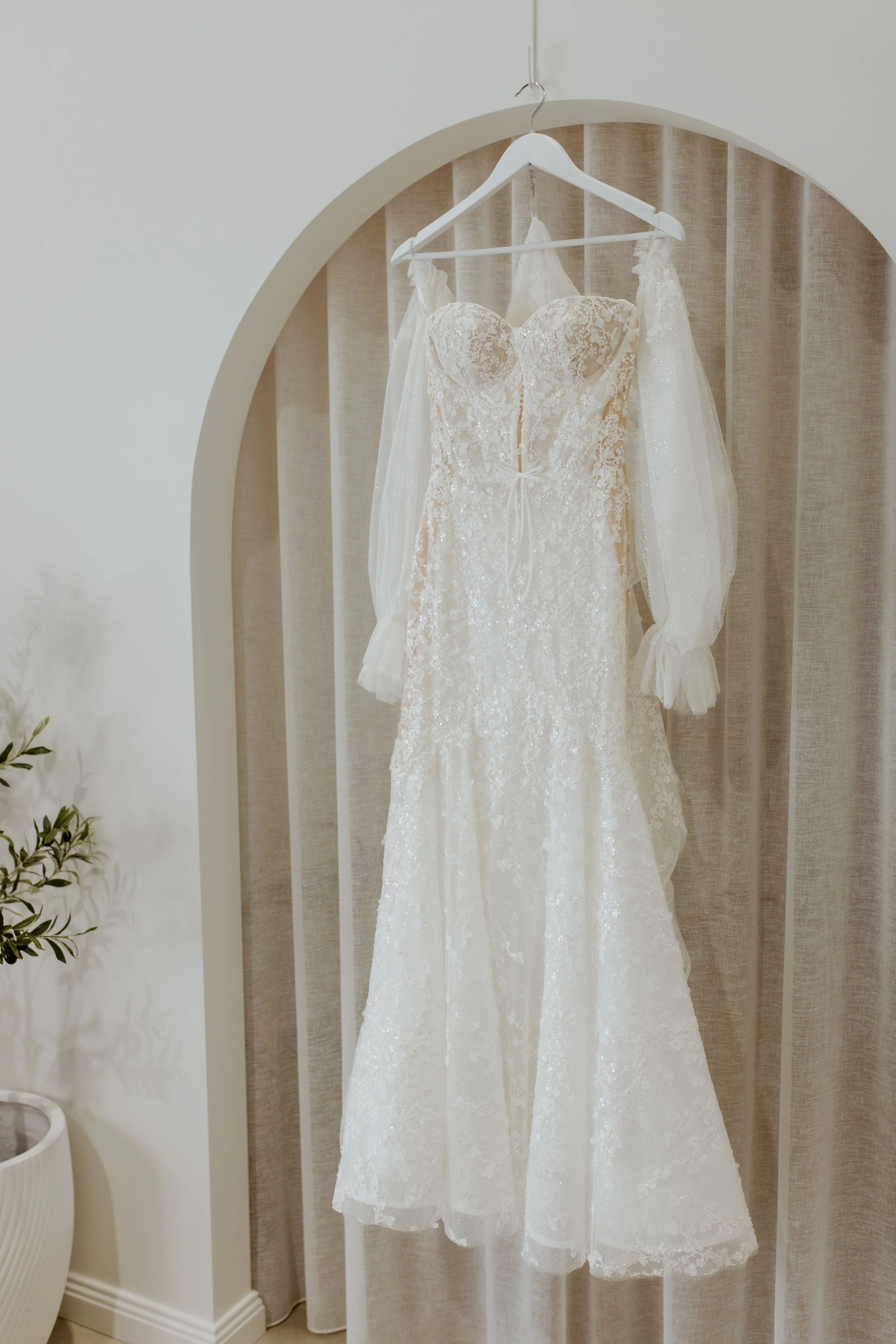 Beautiful white lace wedding dress with sheer long puffed sleeves hanging on a white hanger in front of a beige curtain.