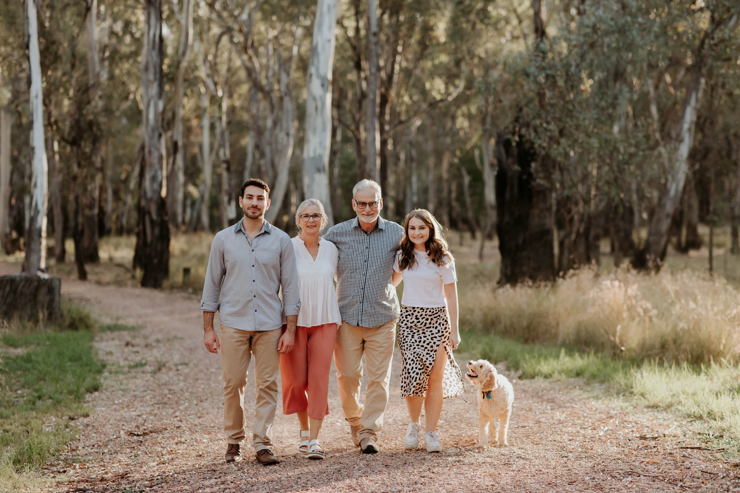 A family of four, including a grandfather, grandmother, son, and granddaughter, walking together on a wooded trail with a small dog, during what appears to be late afternoon or early evening.