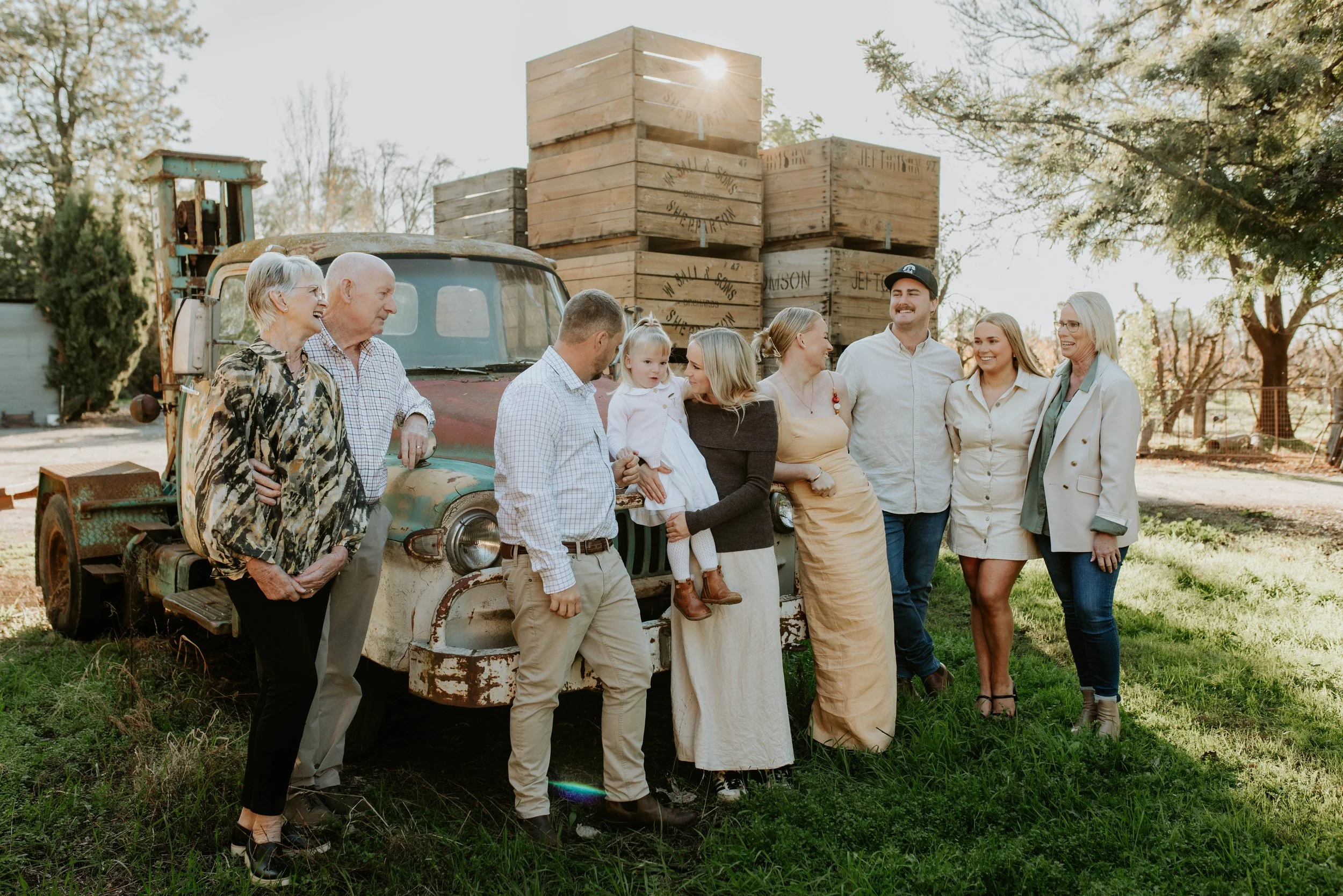 A family group photo outdoors with an old, rusted truck and large wooden crates in the background, during late afternoon sunlight.
