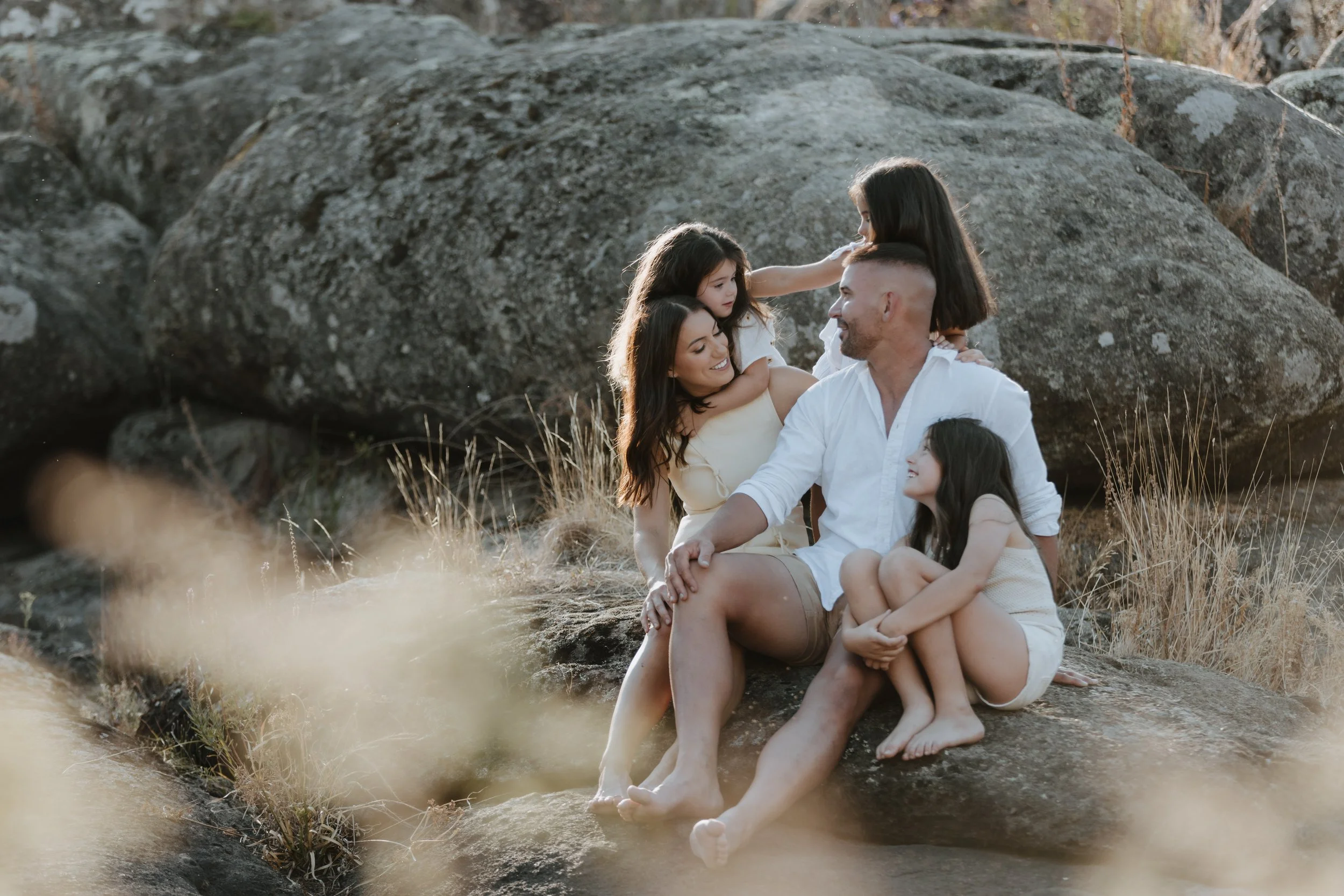 A family of four sitting on a large rock outdoors, surrounded by dry grass and boulders, sharing happy moments and smiles.