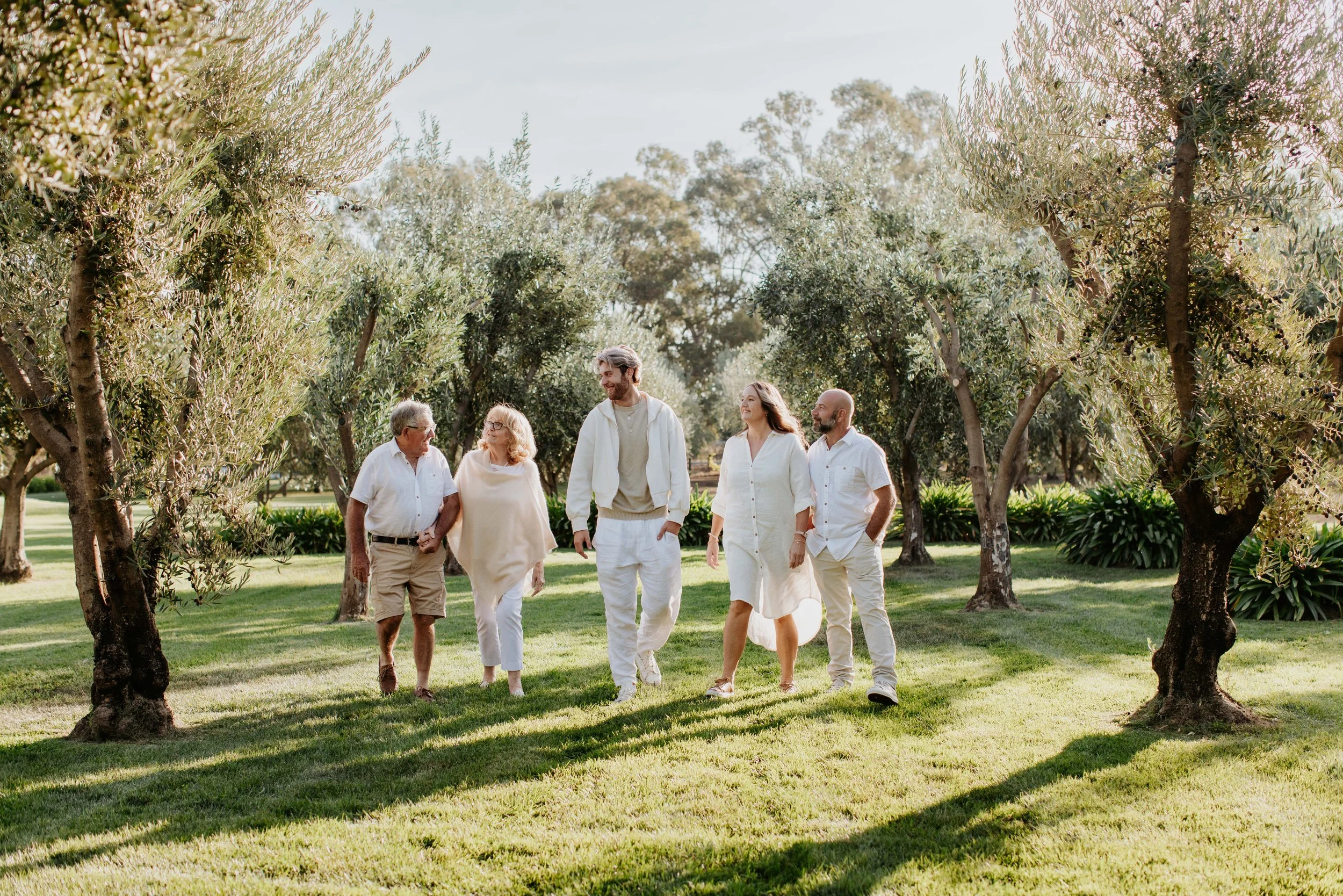 Five people, three men and two women, walking together in a park with green grass and trees, wearing light-colored clothing, enjoying sunlight and smiling. The Owners of the Olivehouse, Kialla.