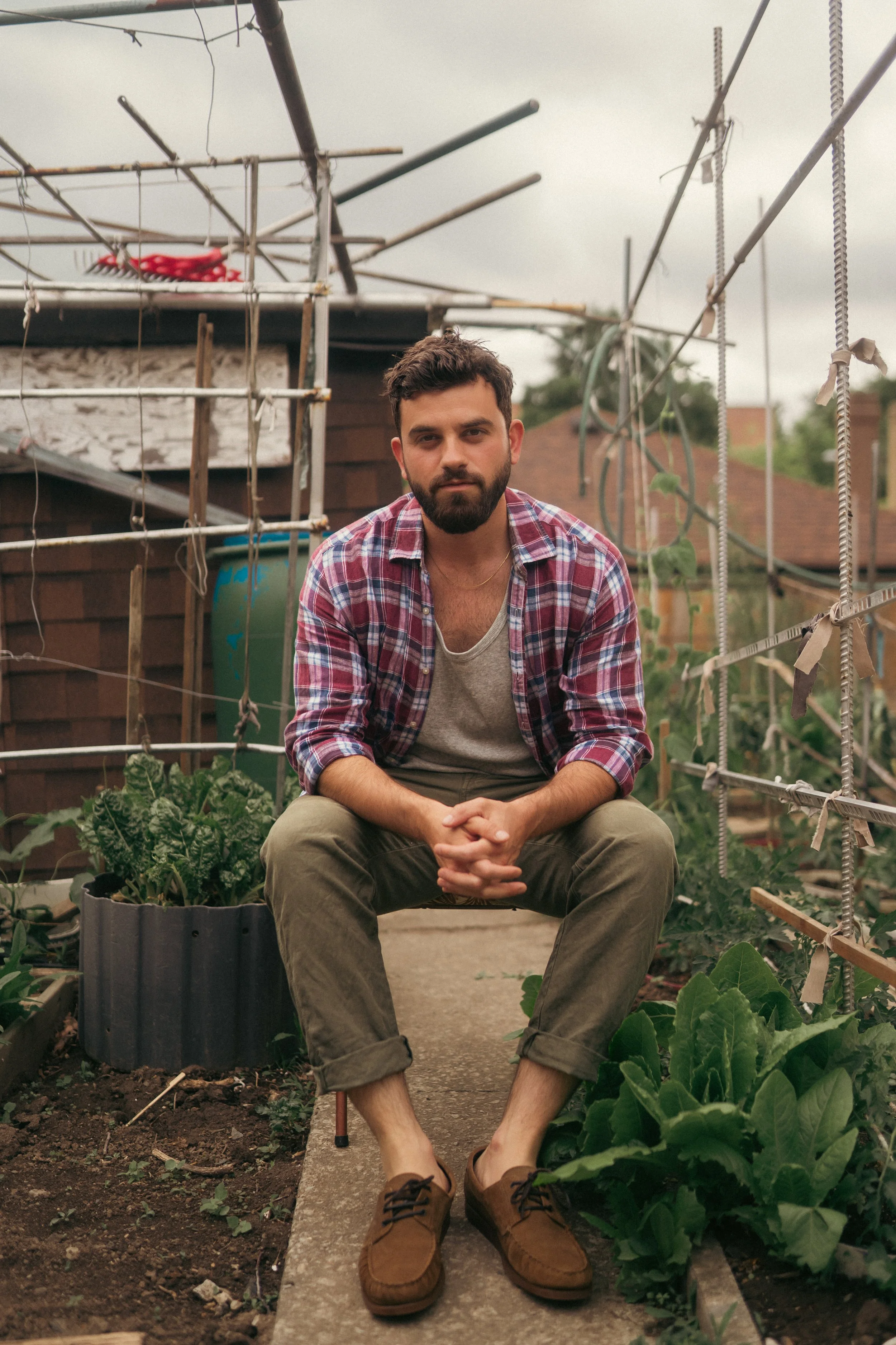 A young man with a beard and short brown hair sits on a concrete path in a garden, surrounded by plants. He wears a red plaid shirt over a gray t-shirt and khaki pants rolled up at the cuffs, with brown shoes. His hands rest on his lap, and he gazes at the camera with a neutral expression. Behind him, there is a garden structure with wooden and metal elements, as well as a green compost bin and various leafy plants.