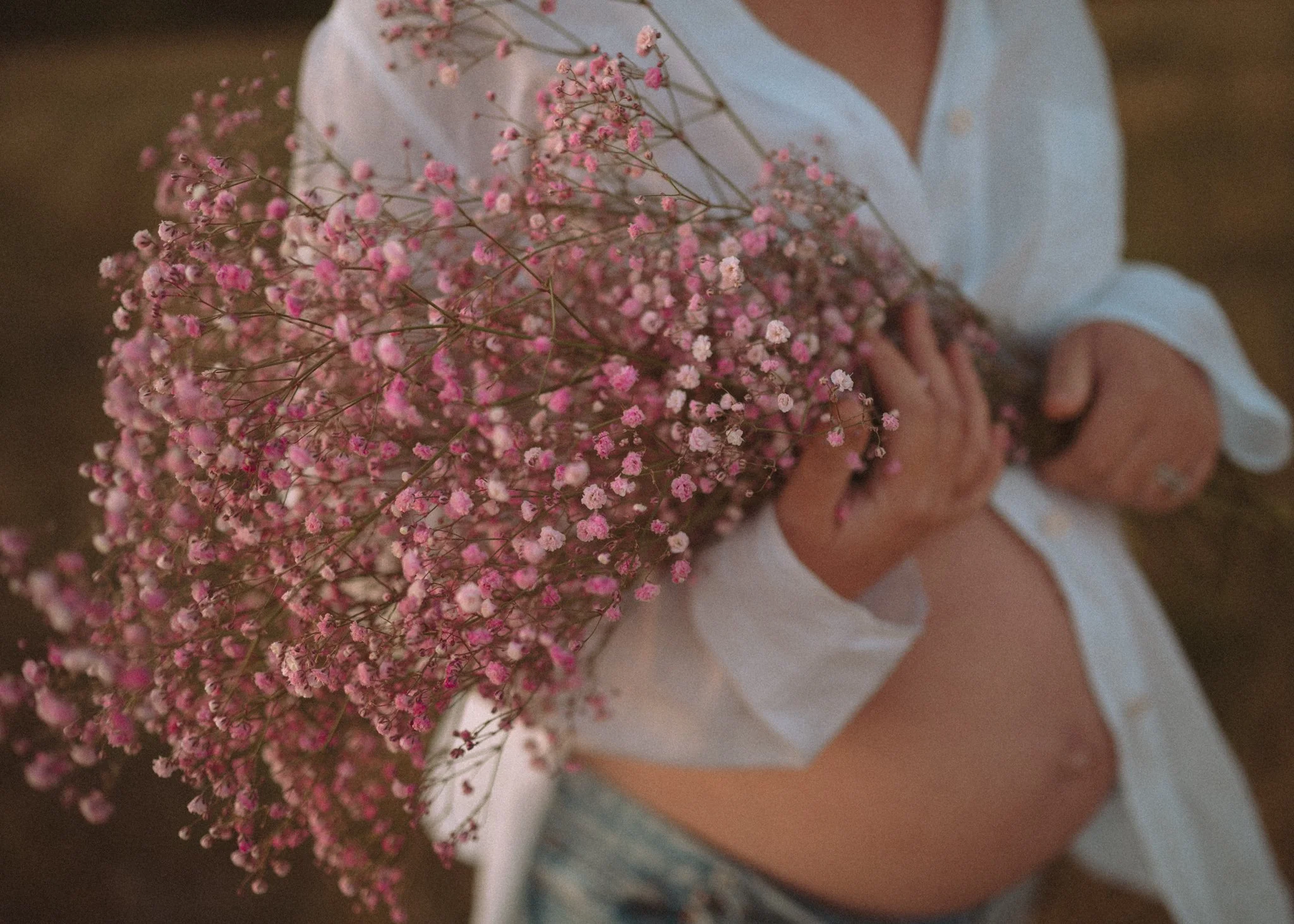 Person sitting outdoors holding a large bouquet of pink baby's breath flowers, wearing a light-colored shirt and denim shorts.
