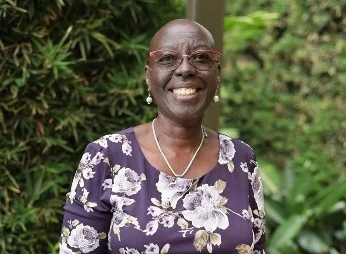 Smiling woman wearing glasses, earrings, and a floral dress outdoors, with green foliage in the background.
