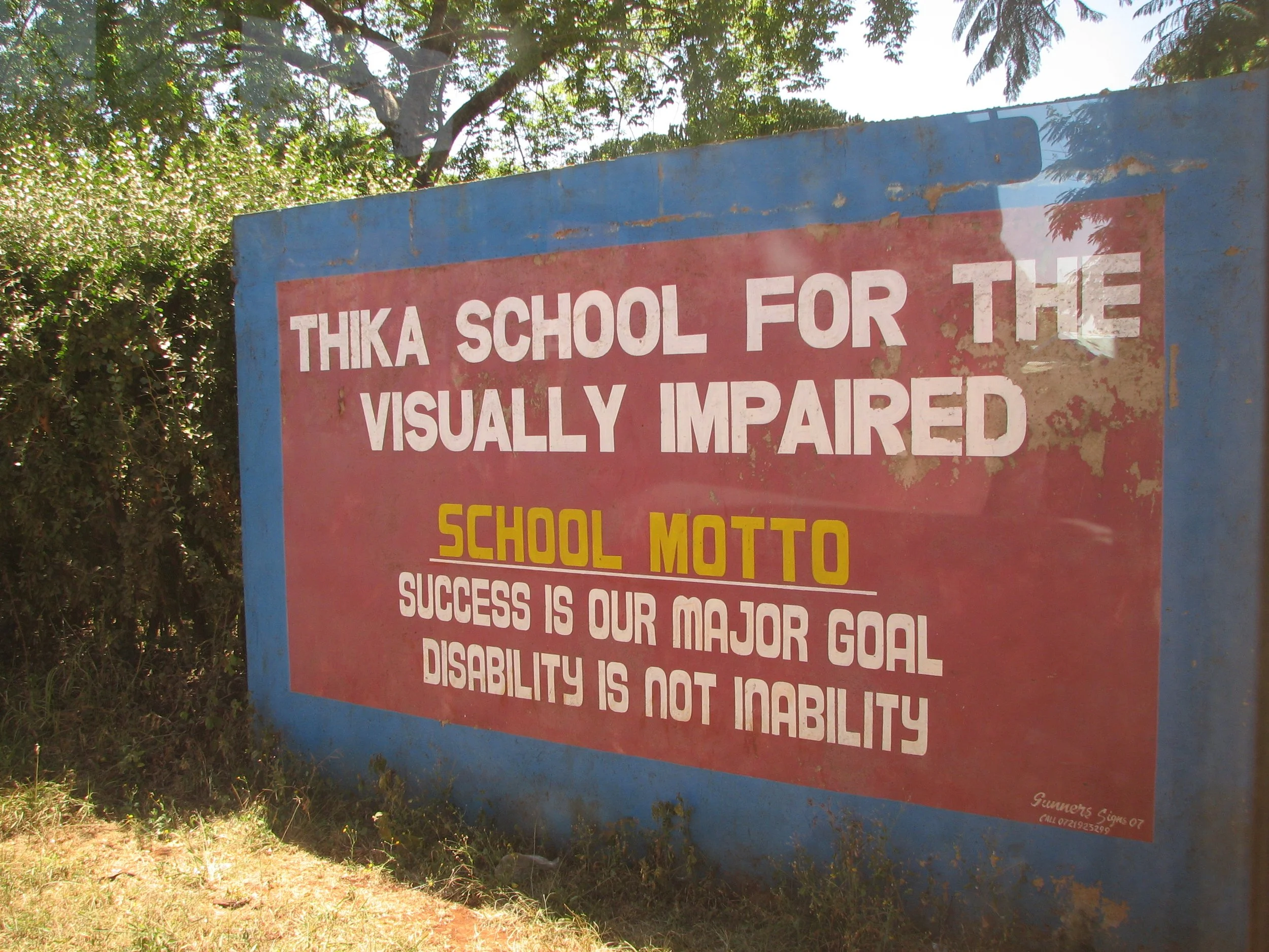 A weathered signboard outdoors with a blue frame and a red background, displaying the words 'THIKA SCHOOL FOR THE VISUALLY IMPAIRED' and additional school motto and success messages, surrounded by greenery and trees.