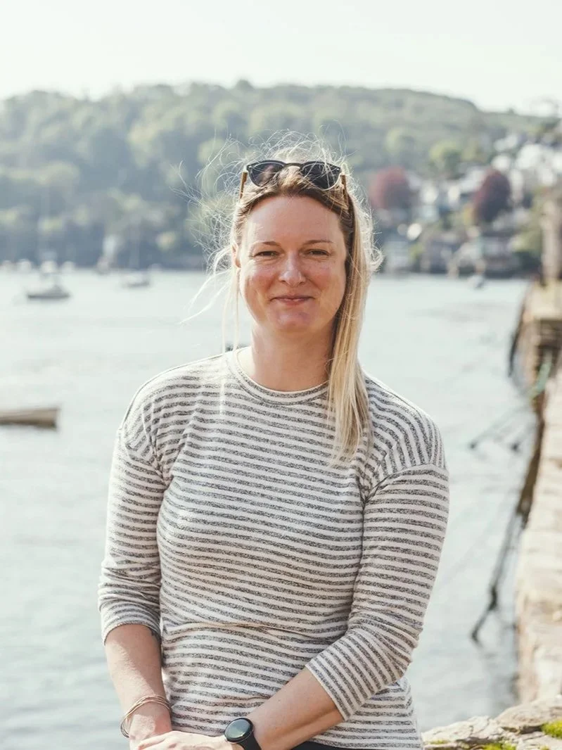 A woman with blonde hair wearing sunglasses on her head and a striped shirt outdoors near a body of water with boats and a hilly landscape in the background.