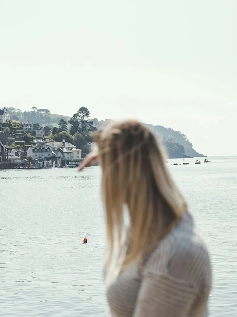 A woman with blonde hair wearing a gray sweater standing near a body of water with boats and houses in the background.