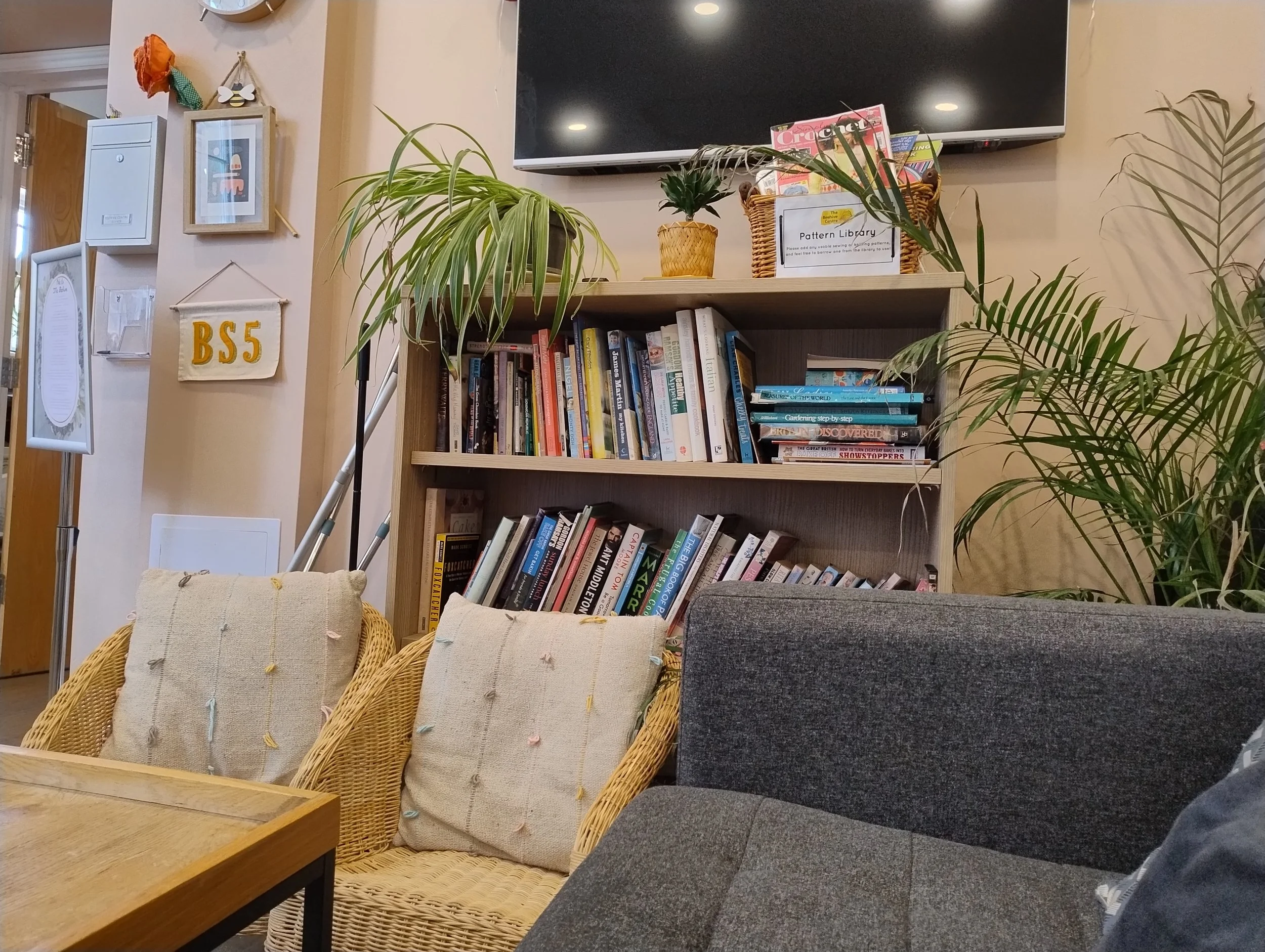 View of a cozy reading corner with a bookshelf filled with books, surrounded by decorative plants, and featuring a wall-mounted TV. Two armchairs with beige cushions and a wooden coffee table are also visible.