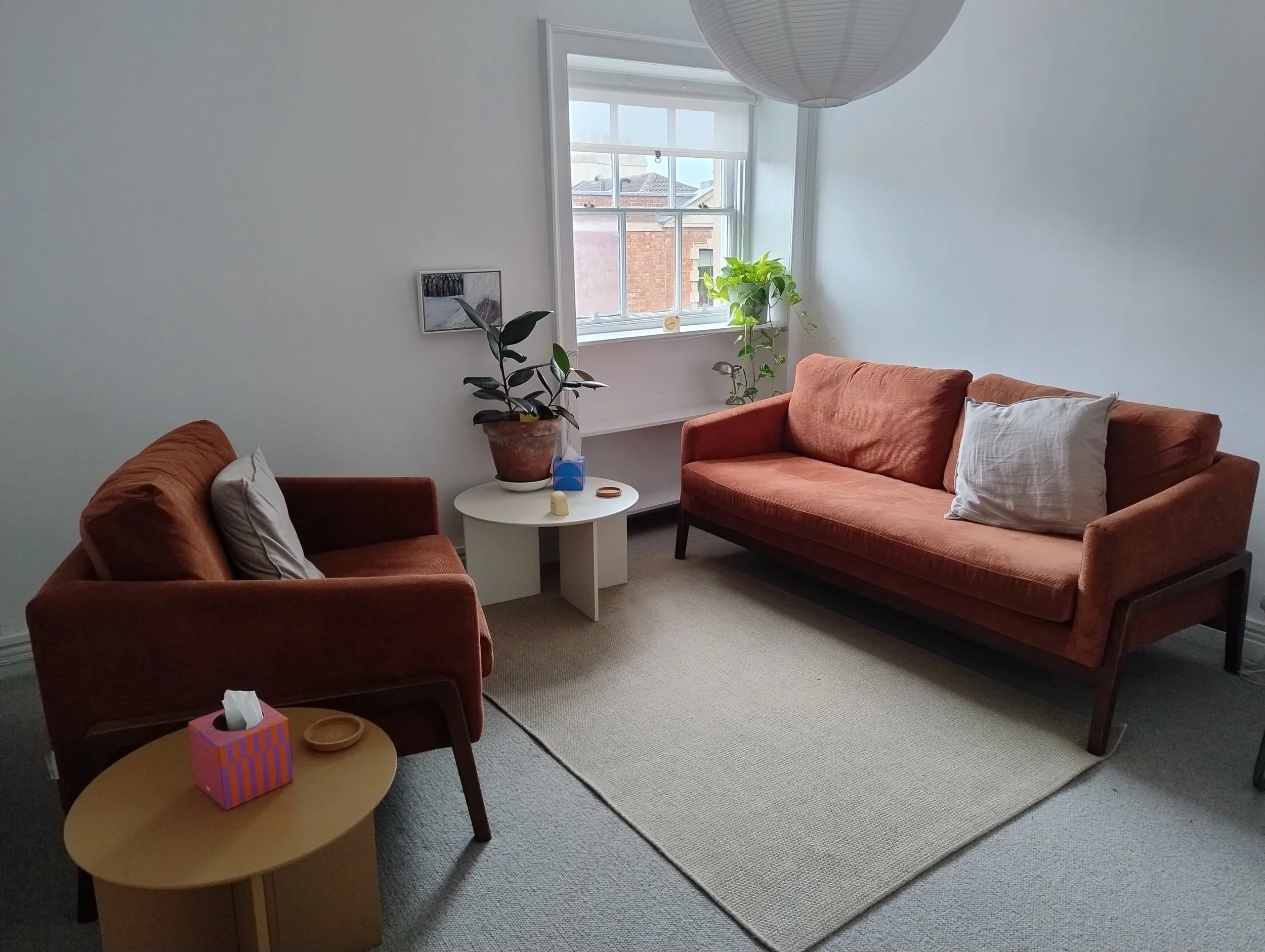 A burnt orange chair faces a sofa of the same colour at a slight angle. There is a window behind the sofa and a plant in the windowsill. A table between to the edge of the room has a plant and coasters.