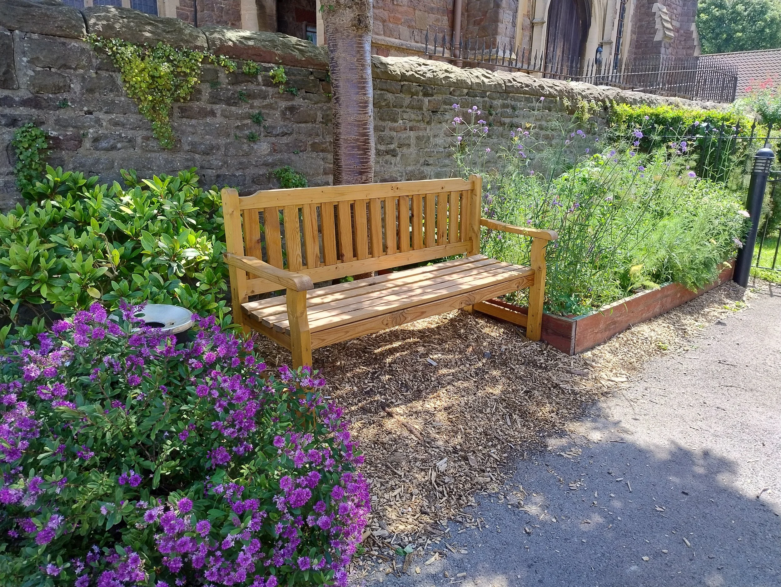 A wooden bench situated beside a flower bed with purple blooms. Behind the flower bed, there is a stone wall with some green plants growing on it, and a tree trunk is visible next to the wall. In the background, there is a building with a fence on top.