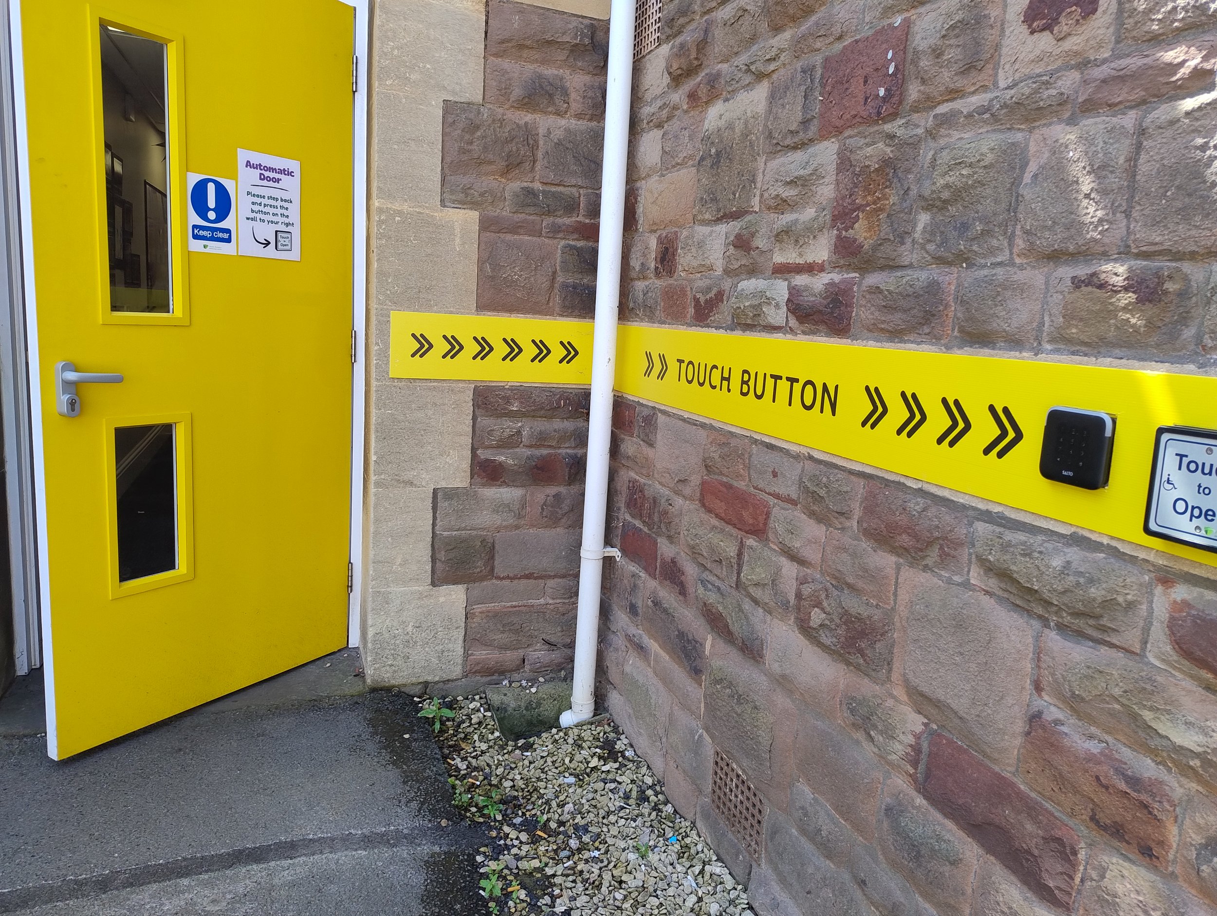 Yellow automatic door with a blue and white sign instructing to keep clear and press a button on the brick wall to open. A yellow tactile band reaches from the door to a 'Touch Button' and a small keypad to open the automatic door.
