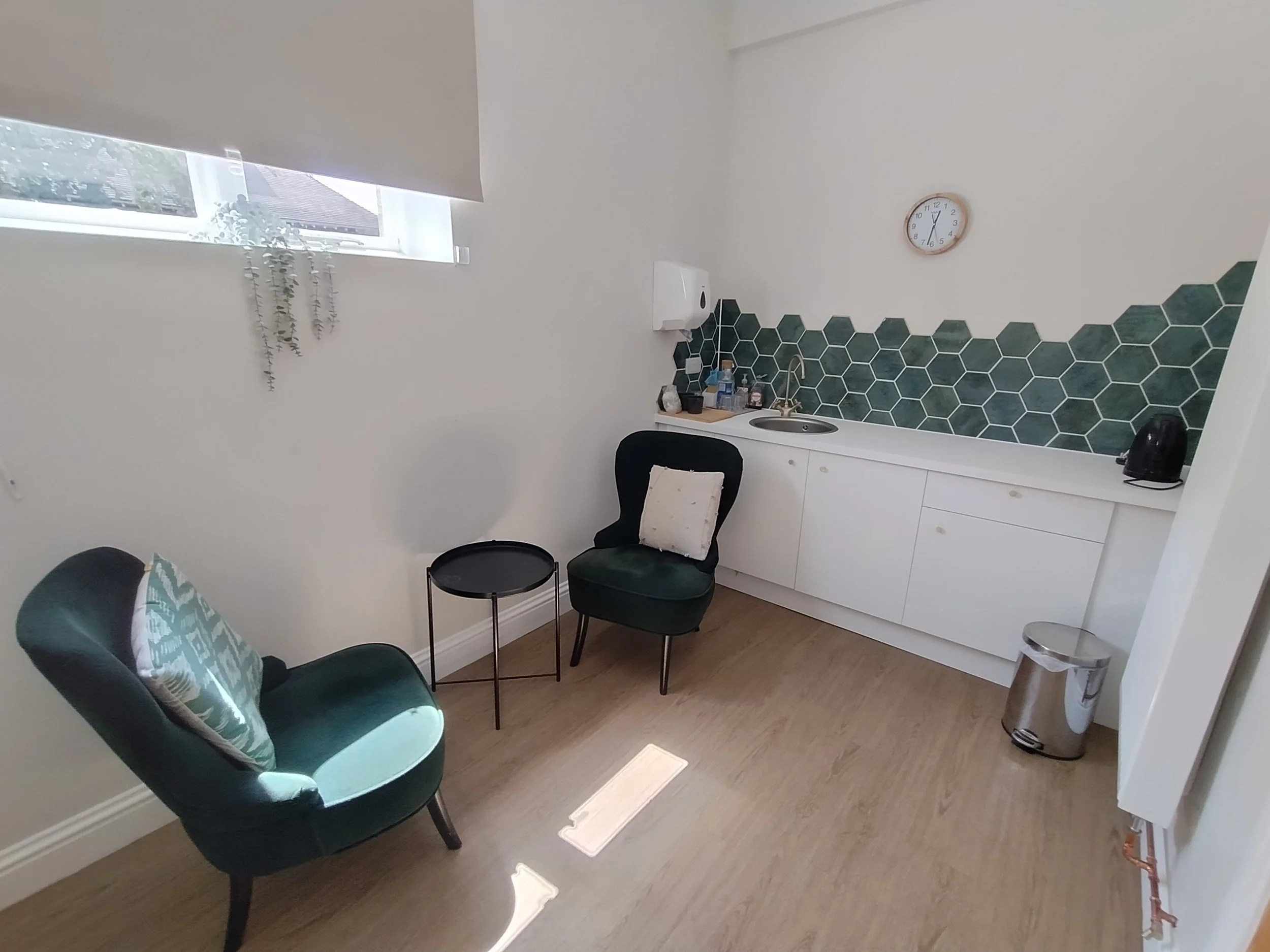A comfortable therapy room with a window, a hanging plant, two chairs facing each other with a low table in between.  Beyond the therapy space is a kitchen counter with a sink and a wall clock.
