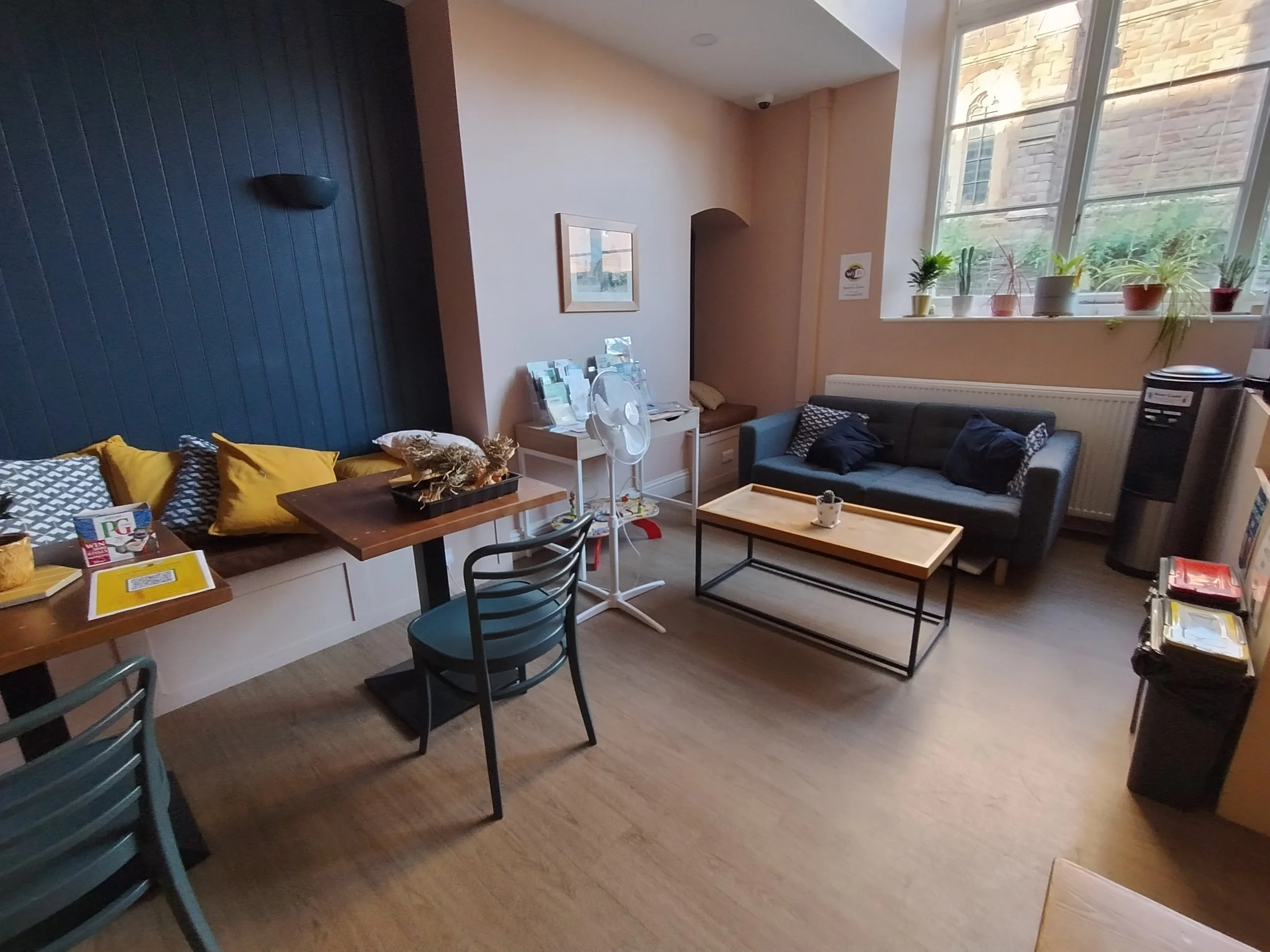 A grey sofa with a low coffee table infront. Cafe style tables are to the left of the room. A large window with plants is behind the sofa. A water dispenser is in the corner with bins and recycling.