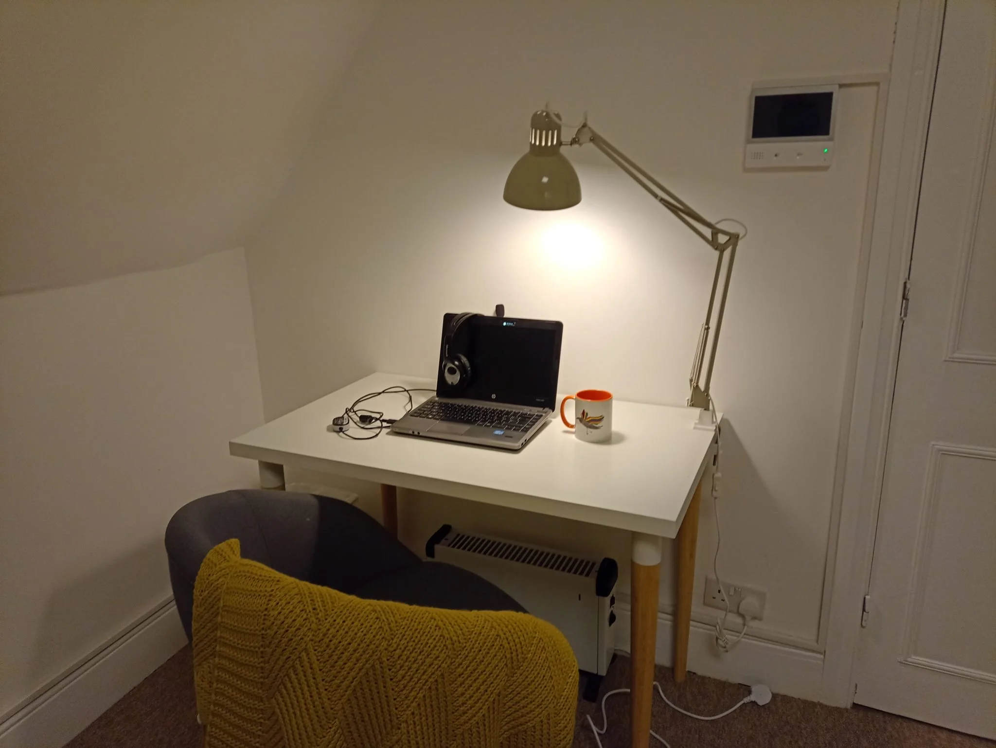 A white desk with a silver laptop, black headphones, and an orange mug on it. A yellow blanket is draped over the grey upholstered desk chair. Above, an adjustable desk lamp is turned on, illuminating the workspace.