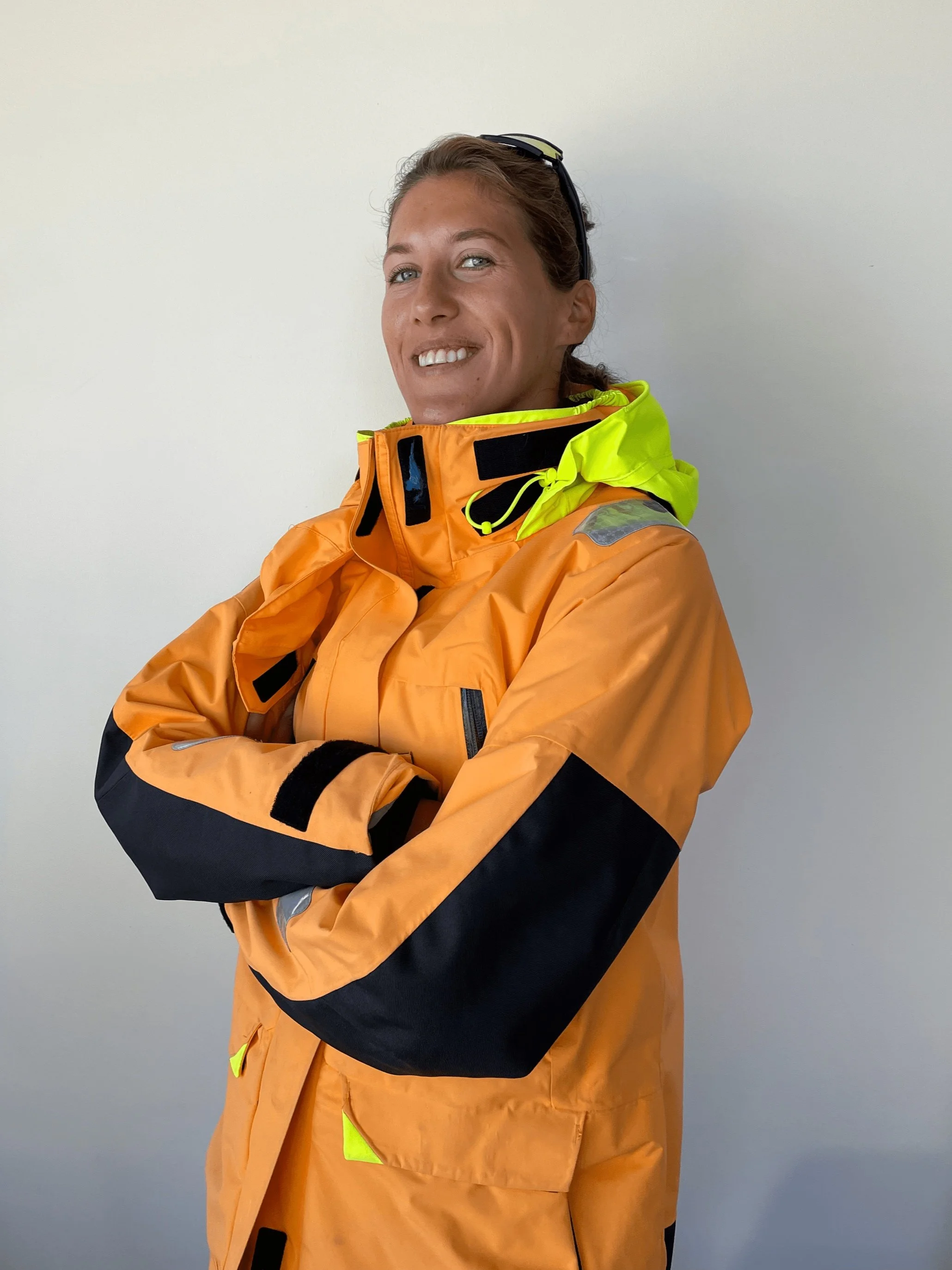 Bild av en kvinne iført oransje og gul varmeklær som smiler, står foran en lys vegg, med armer kryssede.