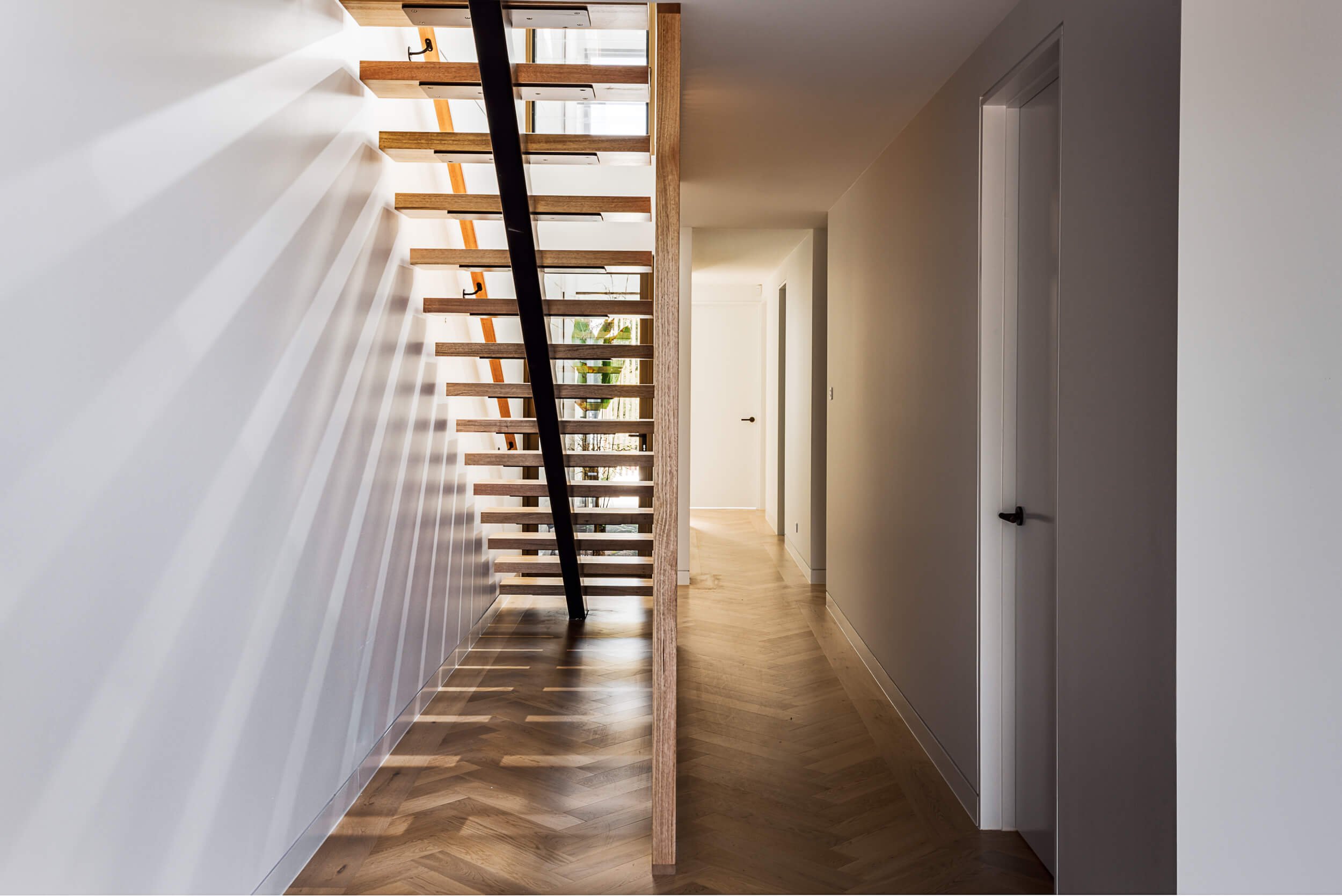 Open-tread timber staircase with black steel spine casting dynamic light patterns across white walls and herringbone timber flooring at O'Brien Duplex Bulli by SLD Projects