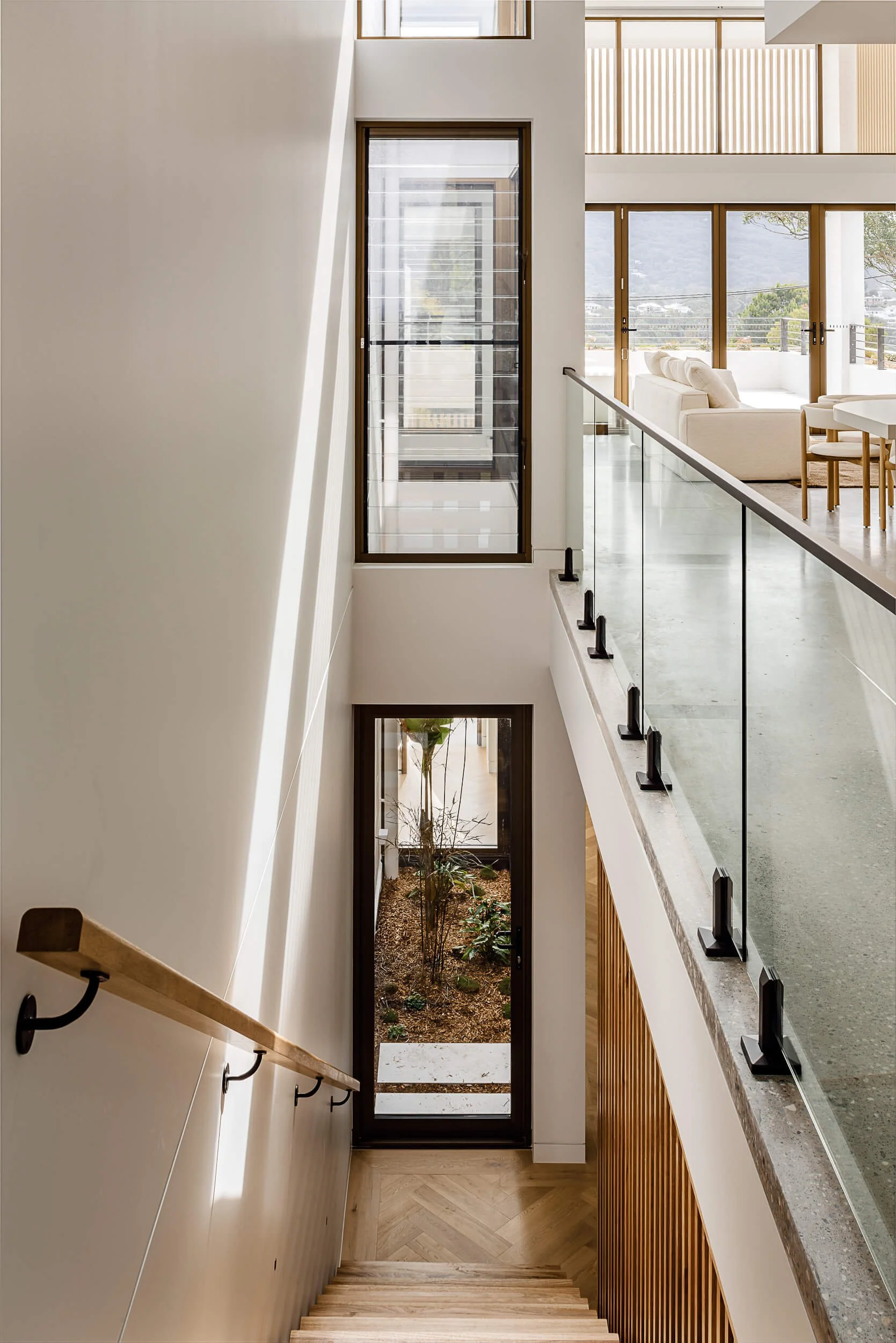 Elevated architectural view down the central light void to the ground floor internal garden, framed by glass balustrades and polished concrete at O'Brien Duplex Bulli by Kibbin Design