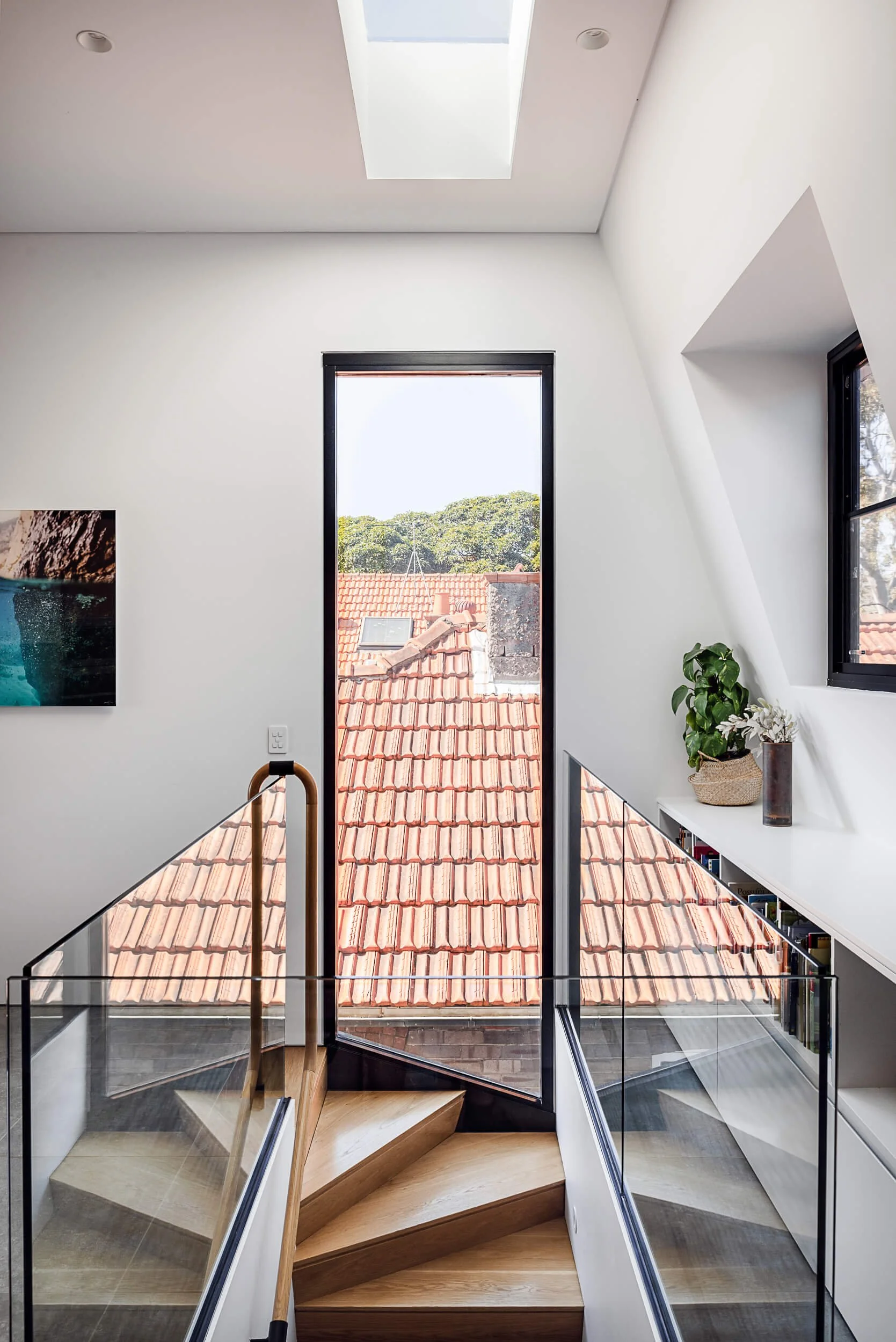 Light-filled timber staircase with a glass balustrade, overhead skylight, and vertical window overlooking terracotta roofs at Bondi Heights in Sydney, NSW