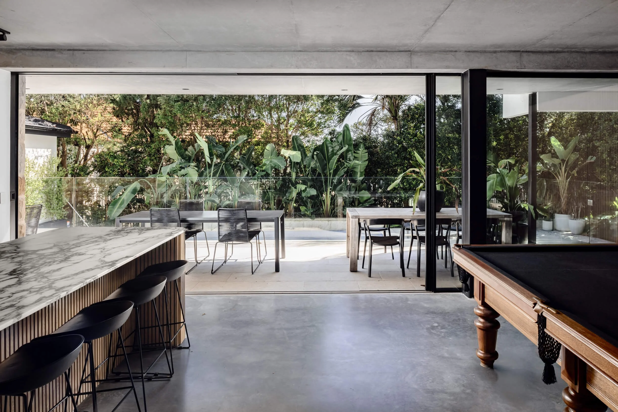 Open-plan living space featuring a marble kitchen island, pool table, polished concrete floors, and open sliding doors to a garden terrace at Stonemasons House in Vaucluse, NSW