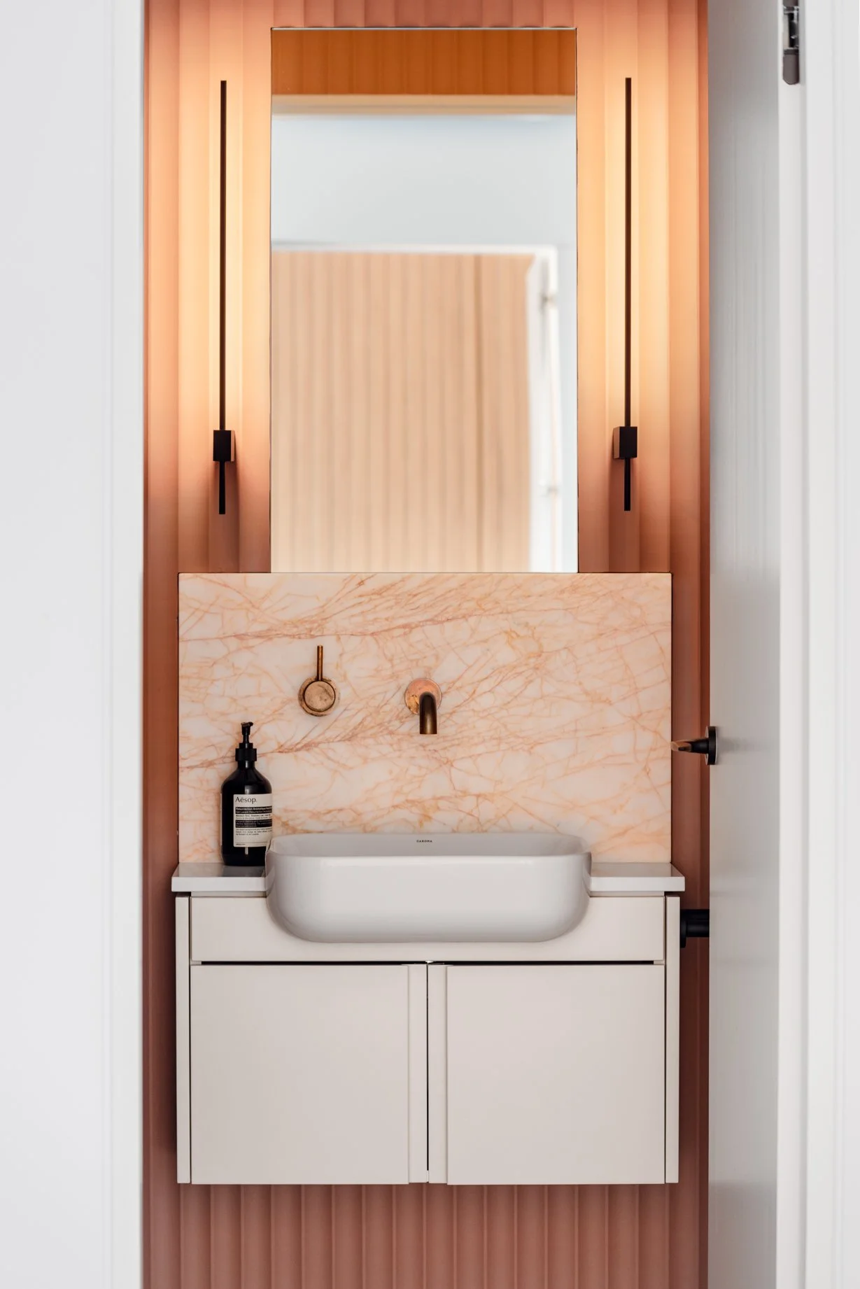 Powder room vanity featuring terracotta fluted wall paneling, veined pink marble splashback, brass tapware, and linear wall sconces at Bondi Heights in Sydney, NSW