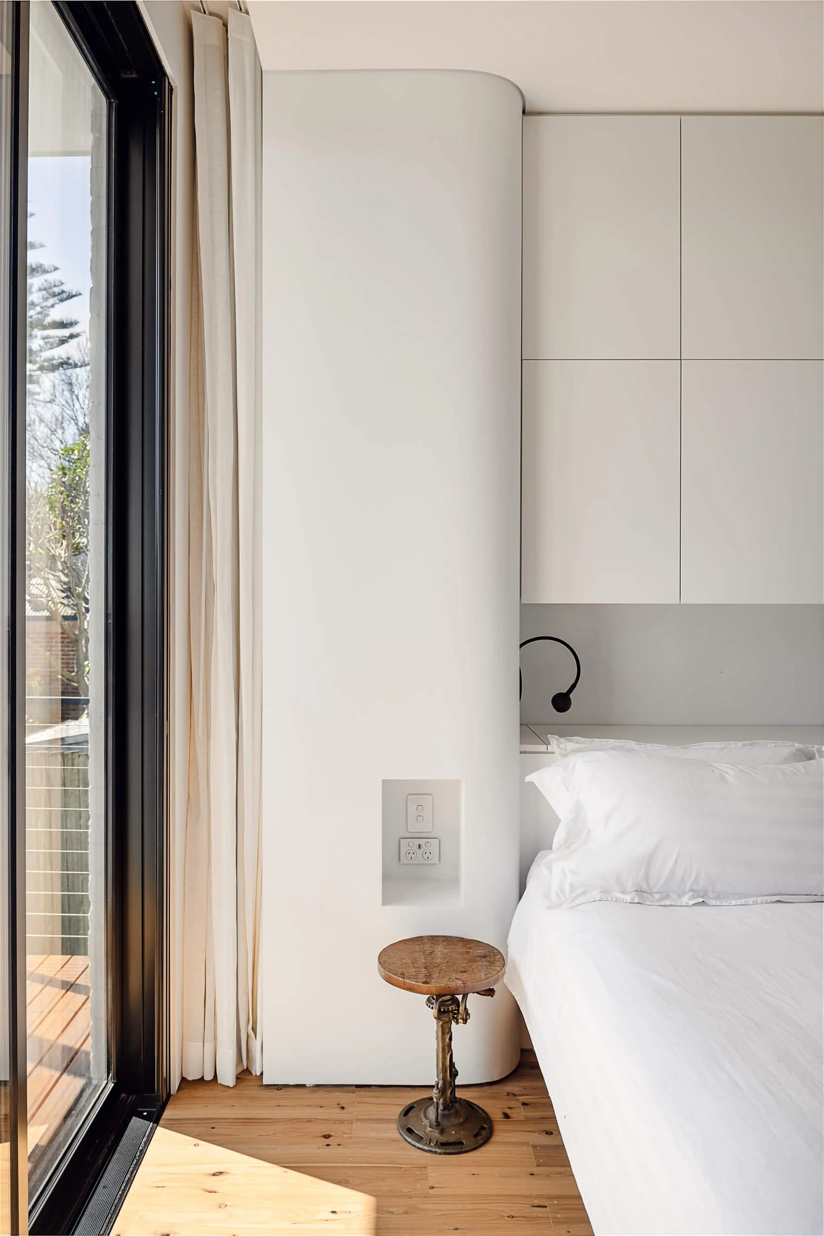 Minimalist bedroom with curved white joinery, built-in bedside niche, and black-framed glass sliding door leading to a timber deck at Bondi Heights in Sydney, NSW