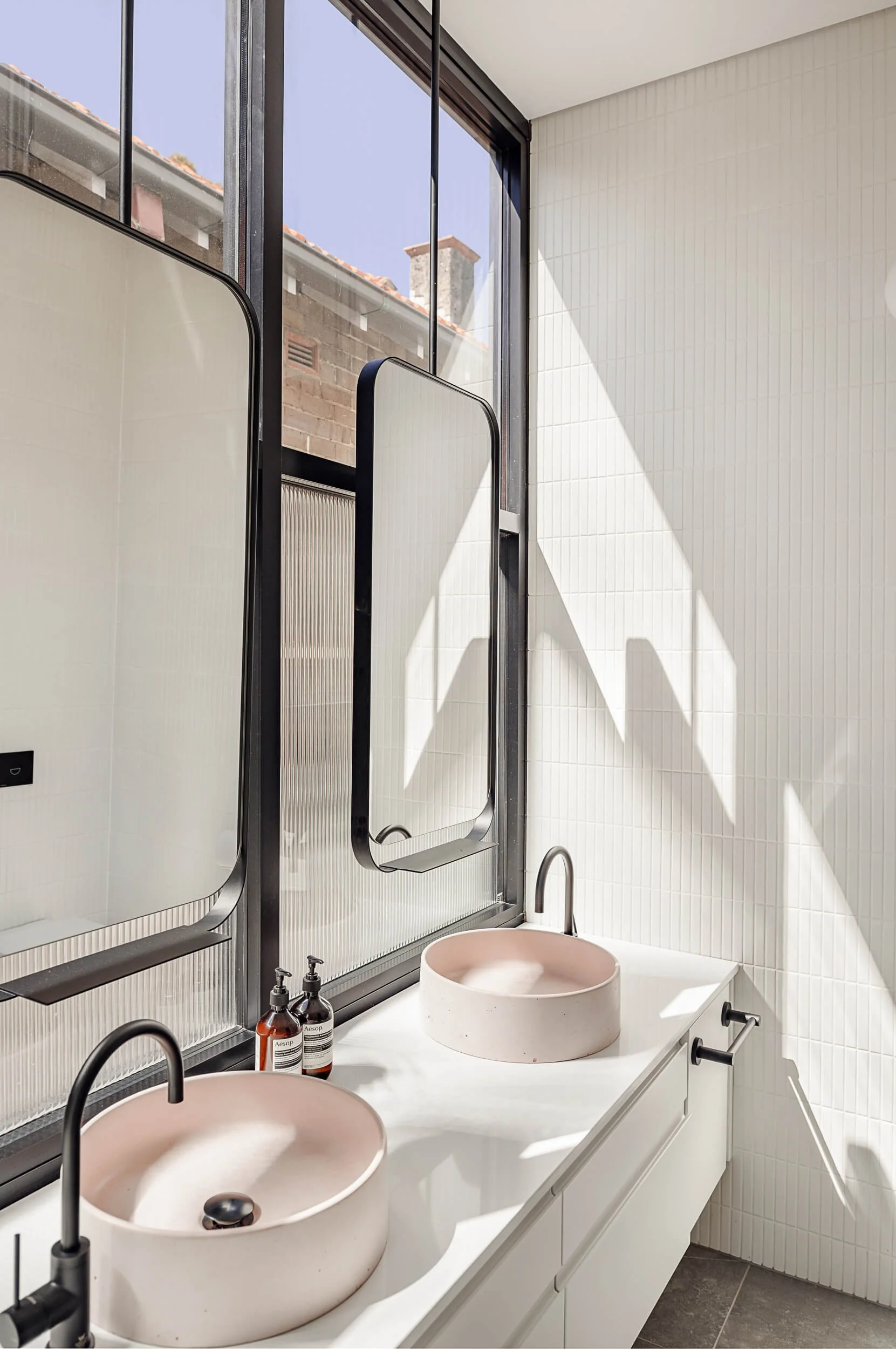 Double bathroom vanity with twin blush pink concrete basins, black-framed mirrors, and white kit-kat tiles with natural light at Bondi Heights in Sydney, NSW