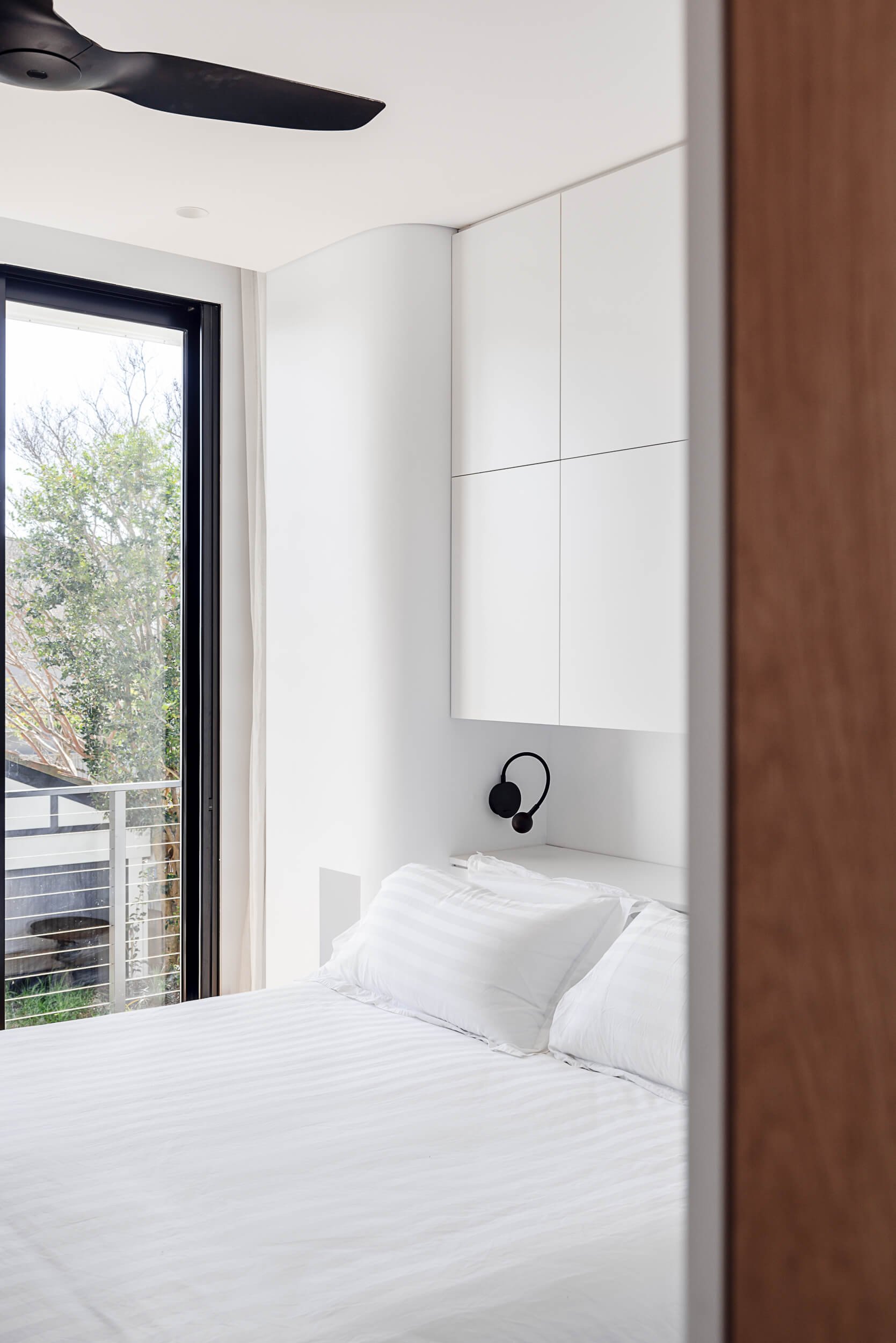Framed view into a light-filled bedroom featuring curved white cabinetry, black wall sconce, and tall window overlooking greenery at Bondi Heights in Sydney, NSW