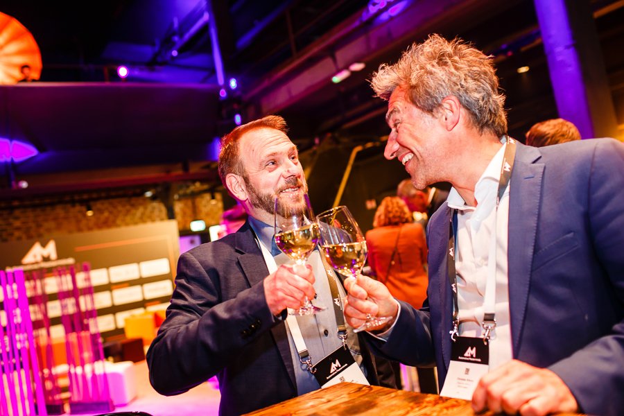 Zwei Männer in Anzügen stoßen mit Wein im Gespräch an, bei einer Veranstaltung in einem modernen, bunten Ambiente.