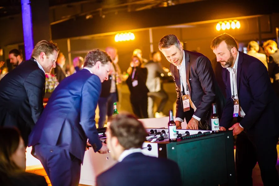 Vier Männer in Anzügen spielen Tischfußball bei einer lustigen Veranstaltung, im Hintergrund weitere Personen in einem festlich beleuchteten Raum.