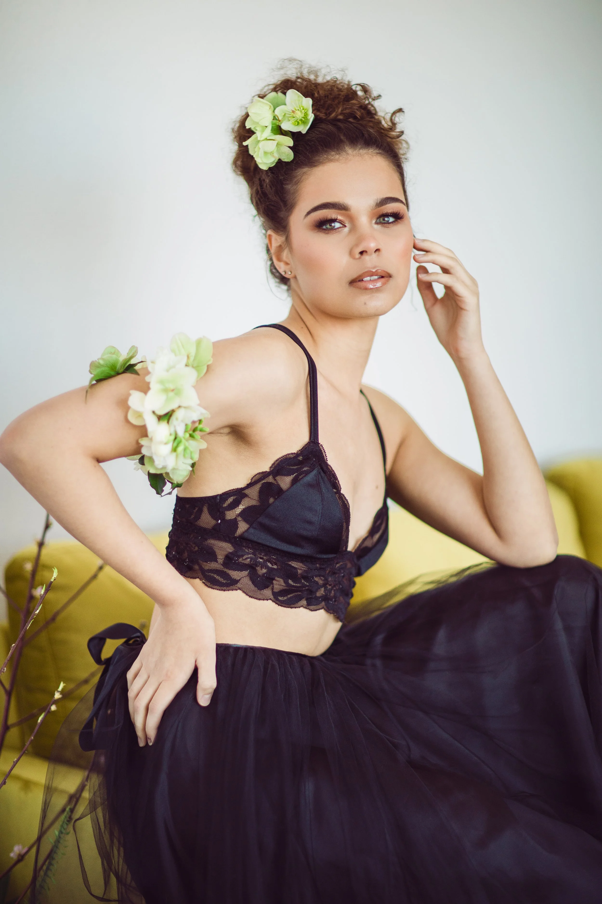 A woman with curly hair and floral accessories, wearing black lace and tulle outfit, sitting on a yellow couch.