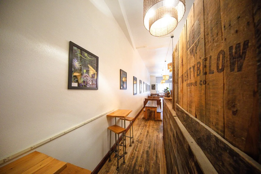 Narrow café interior with wooden tables and benches, framed pictures on the wall, hanging light fixtures, and a large wooden sign that reads "TWENTY BELOW COFFEE CO."