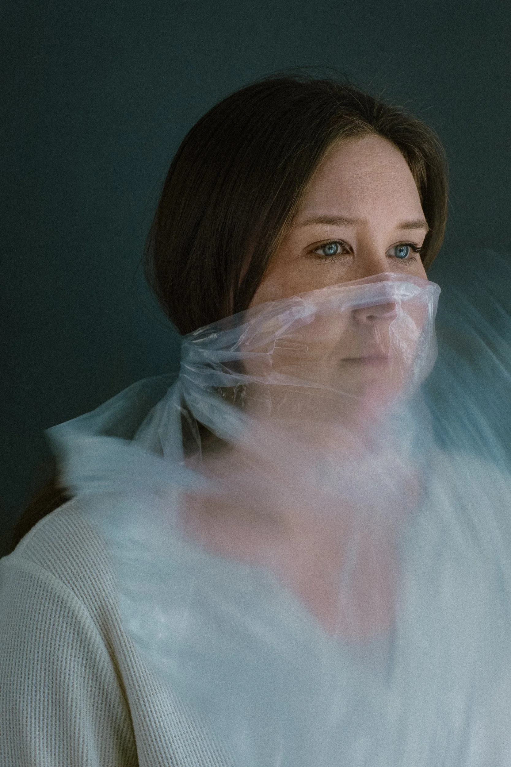 Woman with brown hair and blue eyes wearing a translucent plastic face mask and white shirt, standing against a dark background.