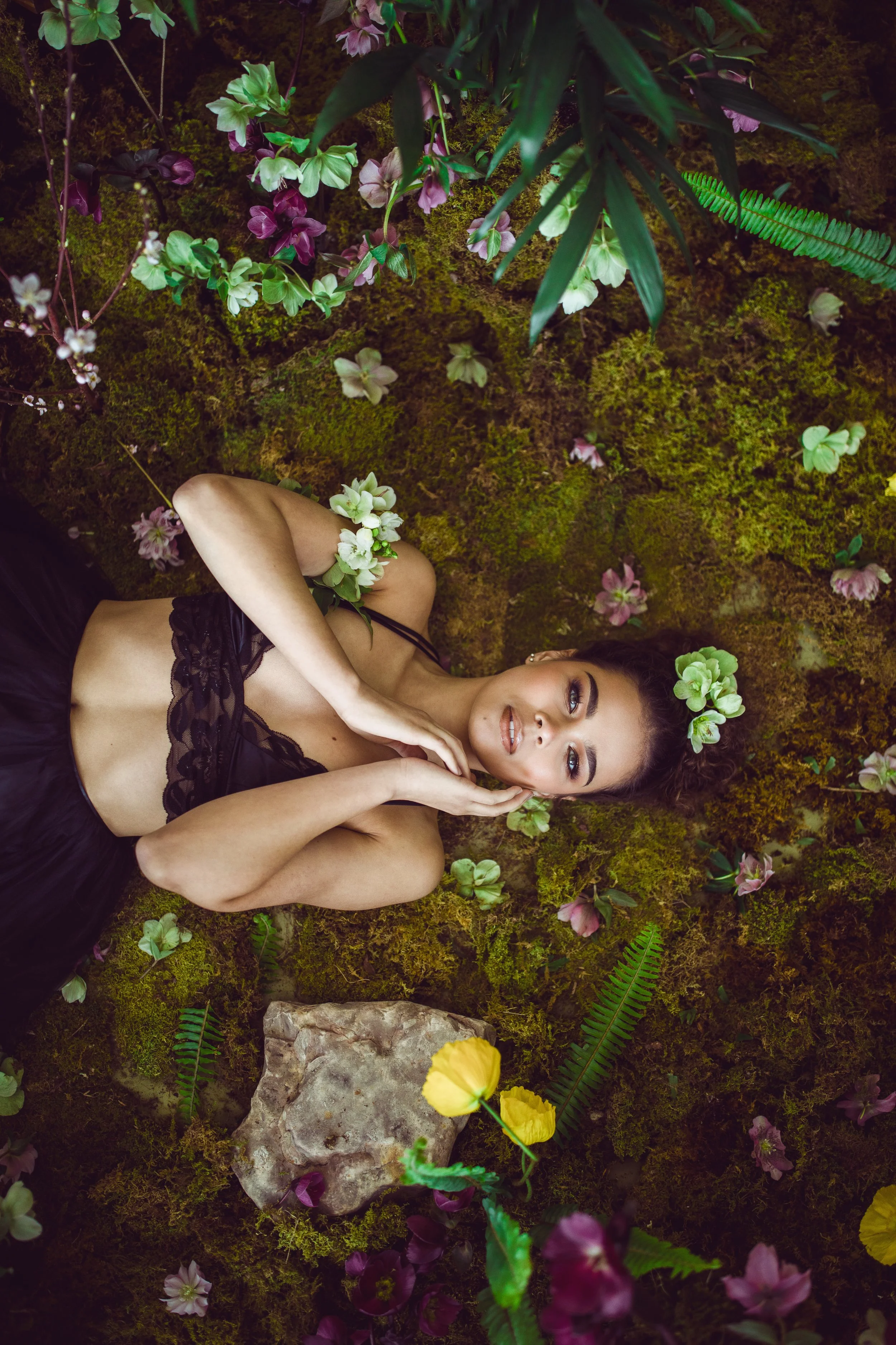 A woman lying on a mossy forest floor surrounded by colorful flowers and green plants, wearing a black lace top with white flowers in her hair.