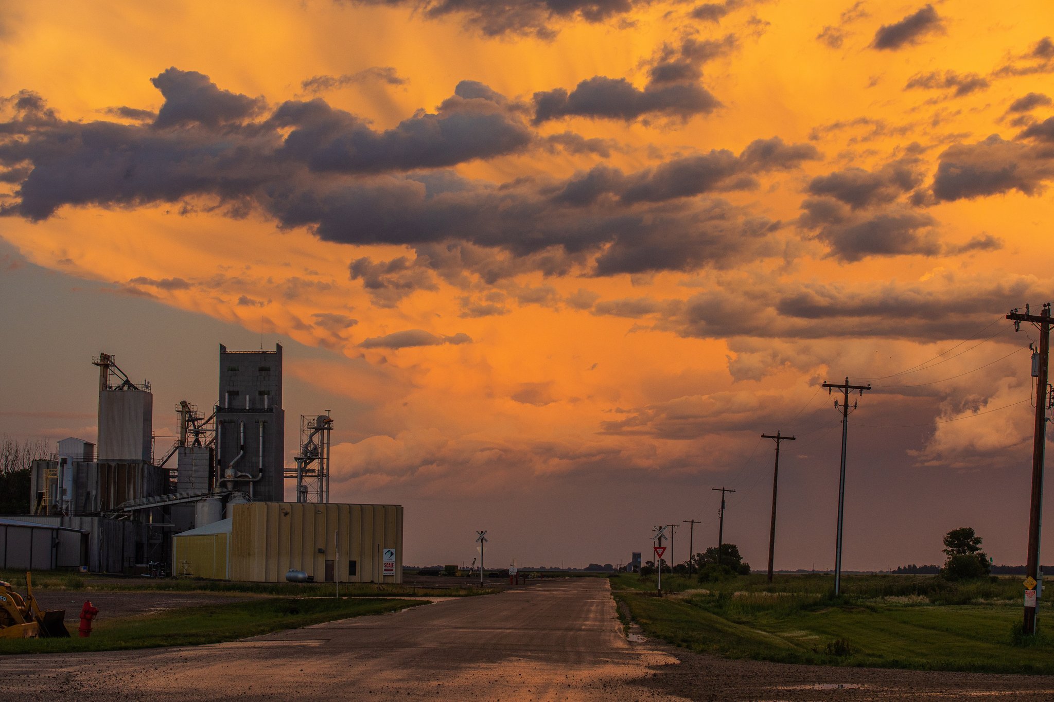 A rural landscape at sunset with a dirt road, utility poles, and a grain elevator against an orange and gray cloudy sky.