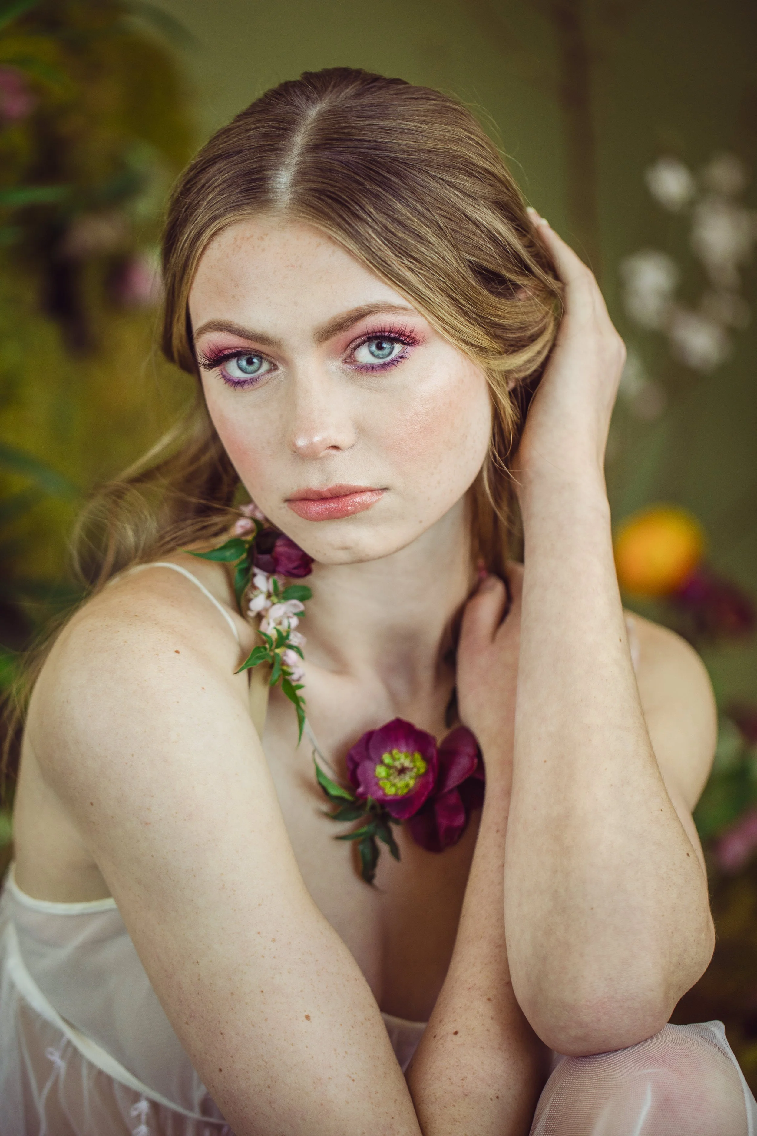 A young woman with fair skin and blue eyes, wearing pink makeup, sitting outdoors surrounded by blurred greenery and flowers. She has long, light brown hair and is touching her head with one hand, while the other rests on her knee. She wears a floral